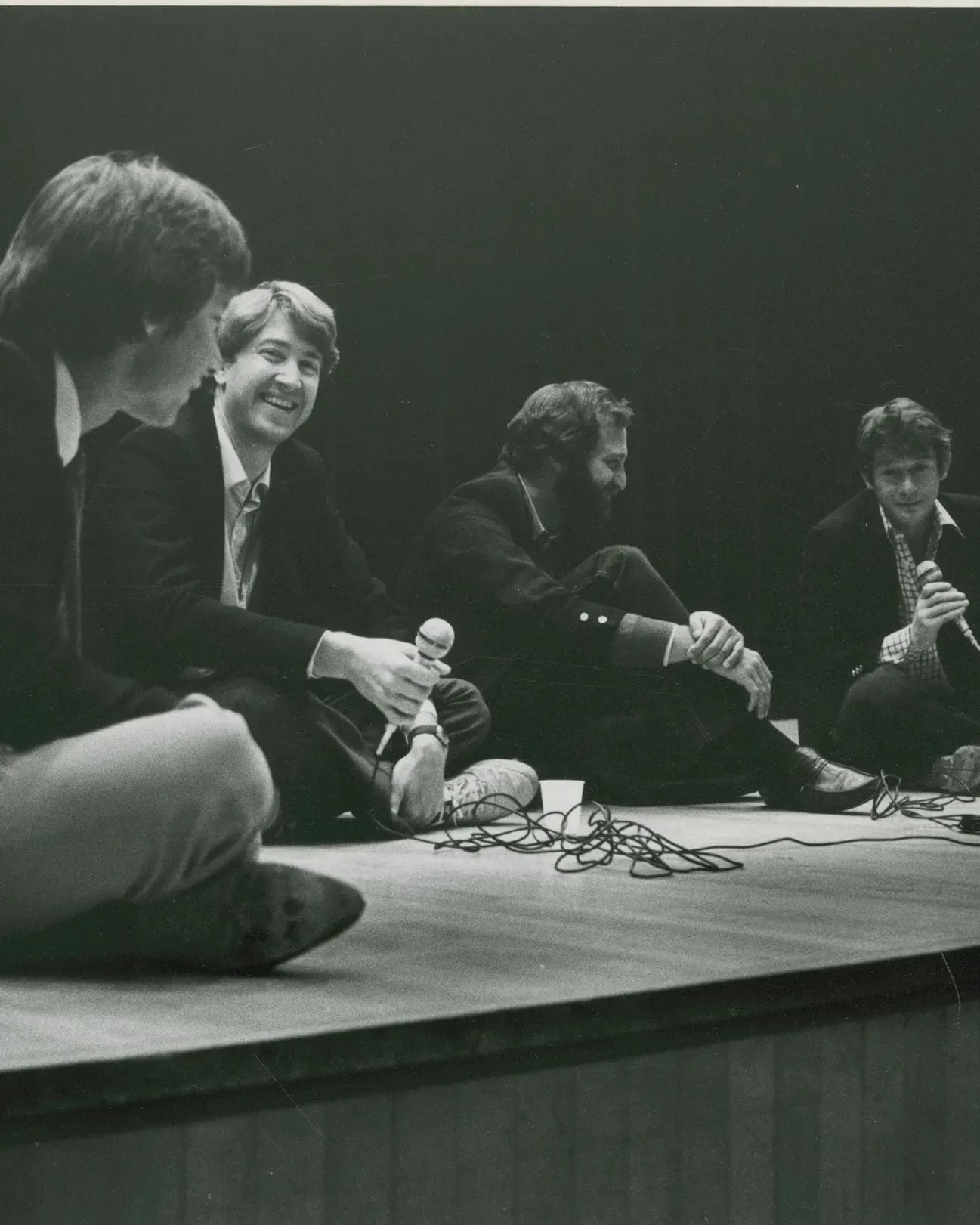 A black-and-white photograph of four men sitting on the floor of a stage, smiling and talking. Two of the men are holding corded microphones.