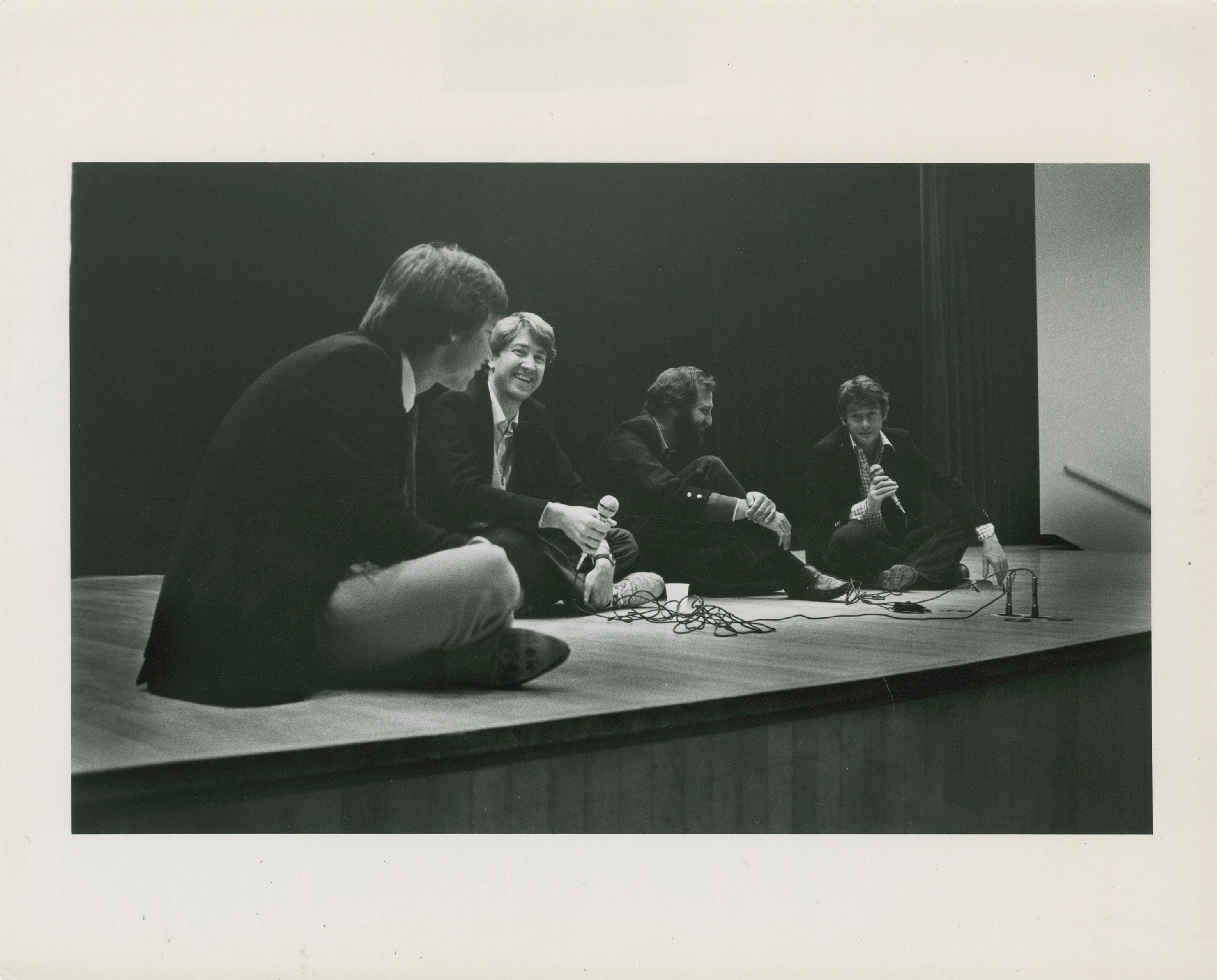 A black-and-white photograph of four men sitting on the floor of a stage, smiling and talking. Two of the men are holding corded microphones.
