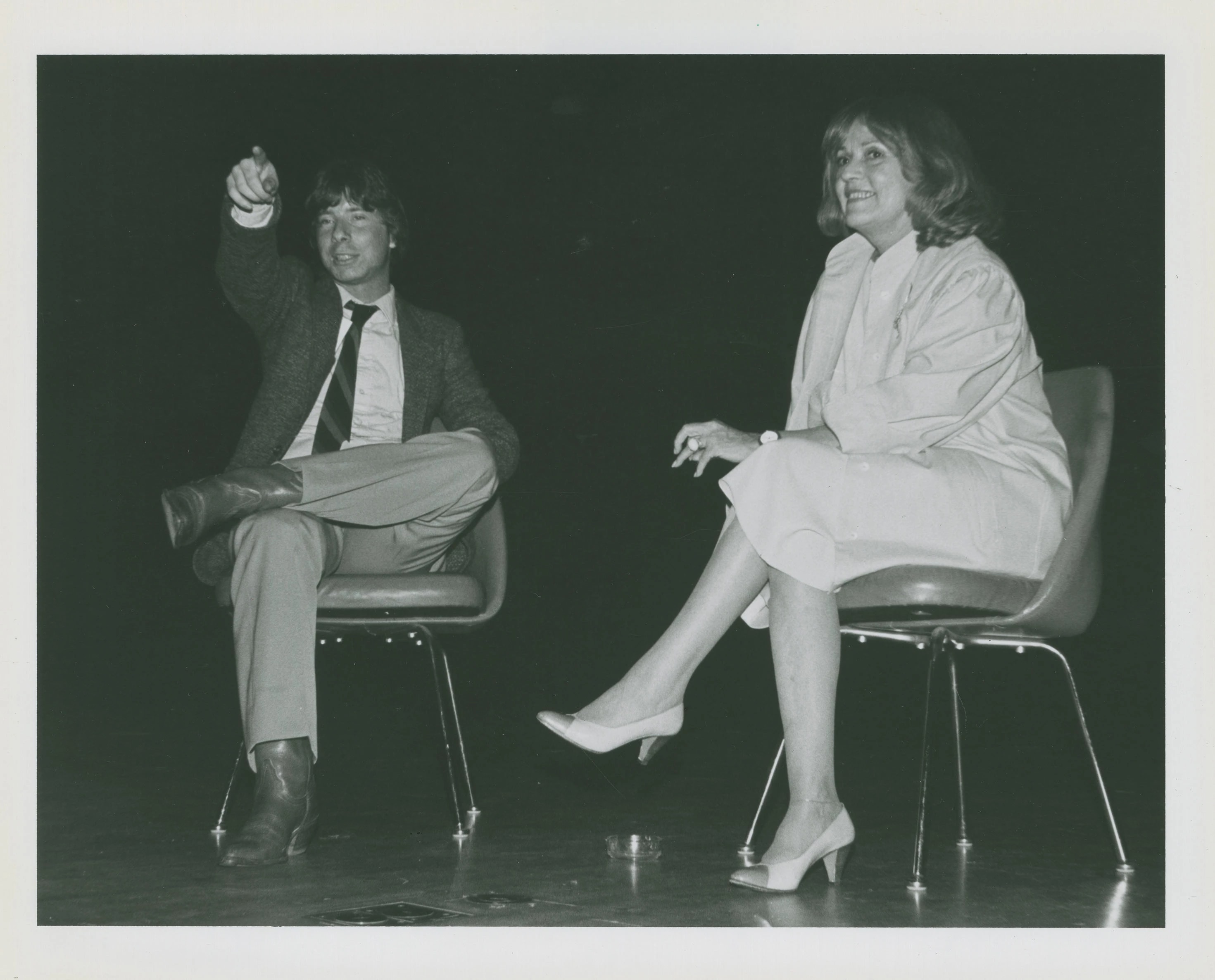 Two people sit in chairs on a stage in mid-discussion. The person on the left is pointing to something off-camera and both people are smiling.