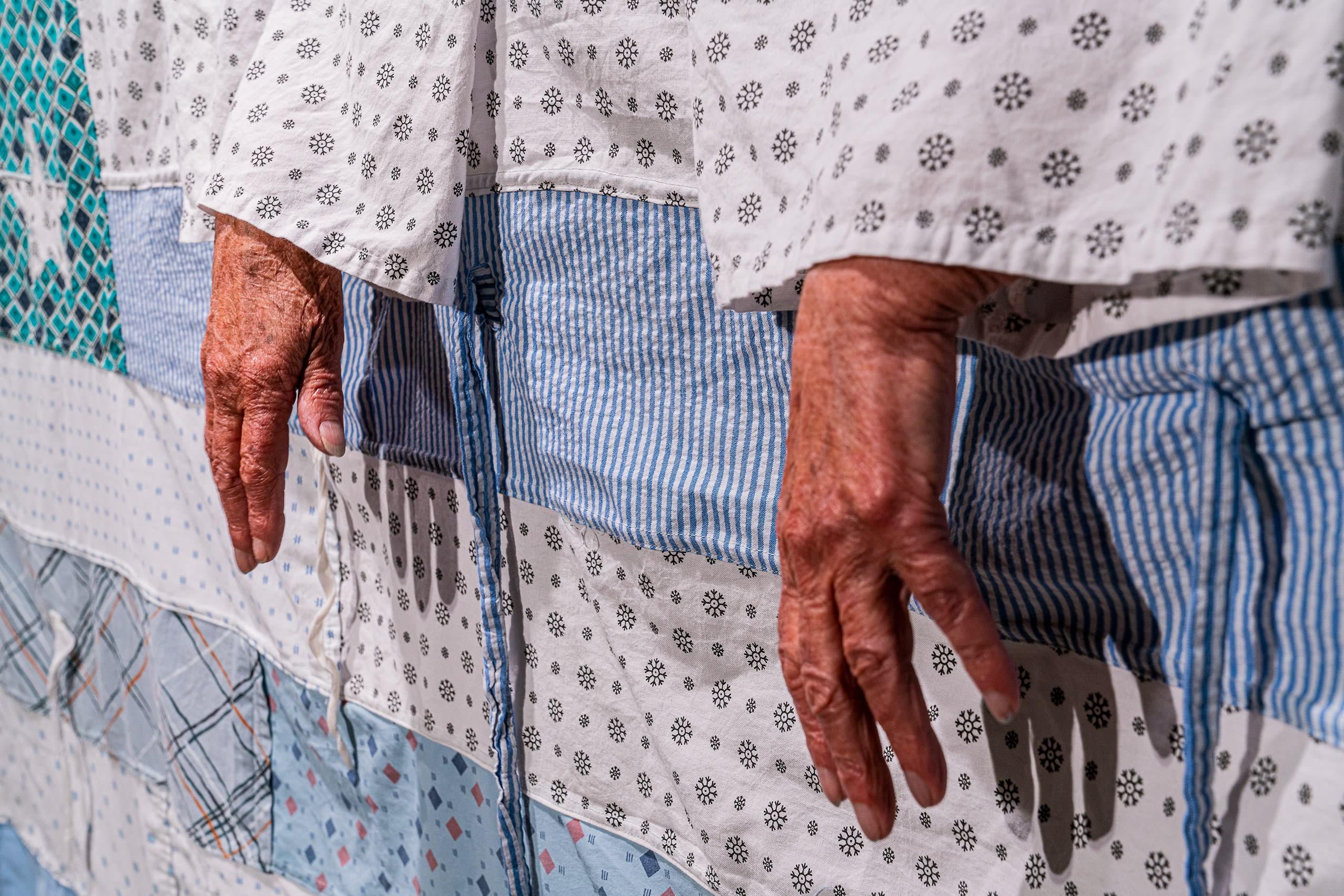 An older person's hands reaching over a patchwork flag made out of blue , white, teal and grey hospital gowns. 