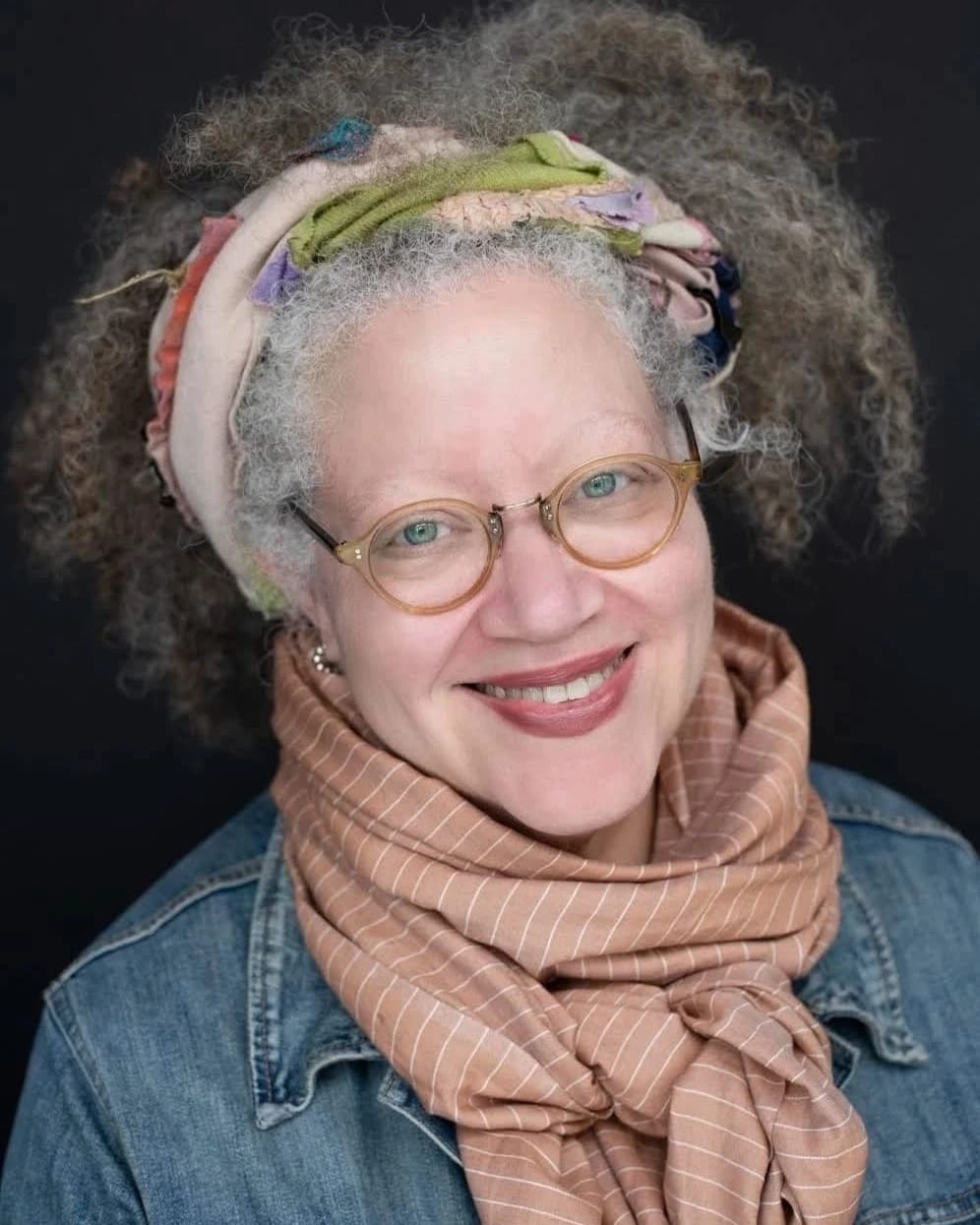 A woman with a bandana in her gray hair smiles, wearing a jean jacket and a colorful scarf.