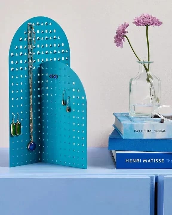A blue jewelry stand made up of two arch shaped planes on a light blue table. There's also blue books and two flowers in a small glass vase.