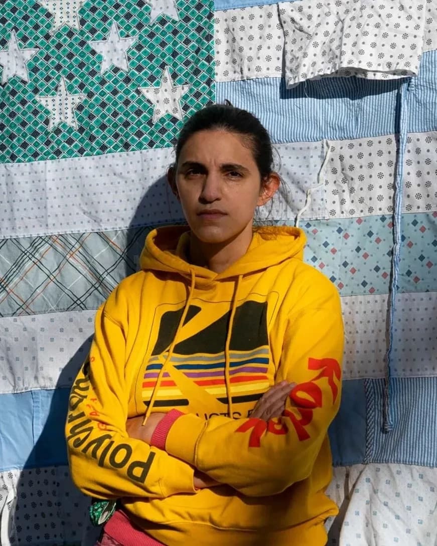 A person in a bright yellow hoodie stands against a quilt resembling an American flag. The quilt has blue and patterned stars and stripes. The mood is calm and contemplative.
