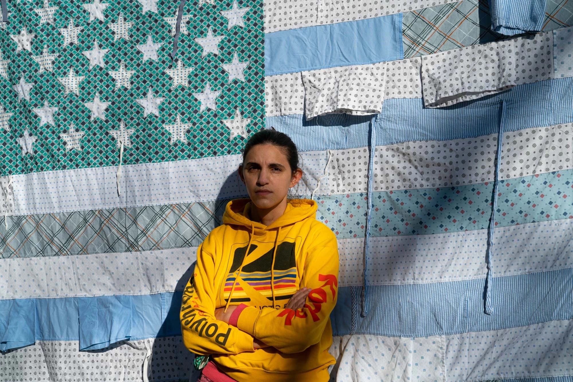 A person in a bright yellow hoodie stands against a quilt resembling an American flag. The quilt has blue and patterned stars and stripes. The mood is calm and contemplative.