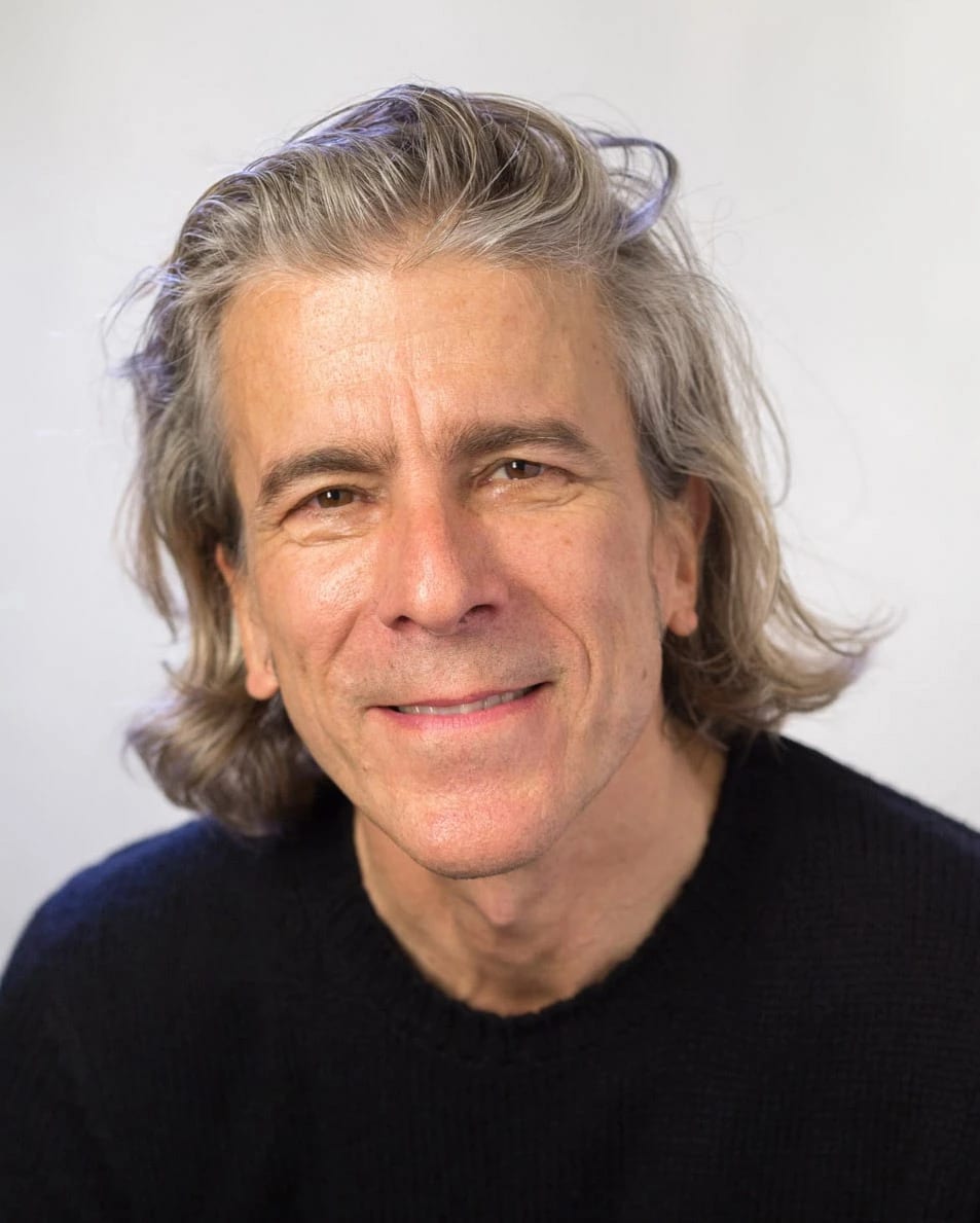 A photo of an older man with gray hair that goes past his ears wearing a black shirt and smiling at the camera.