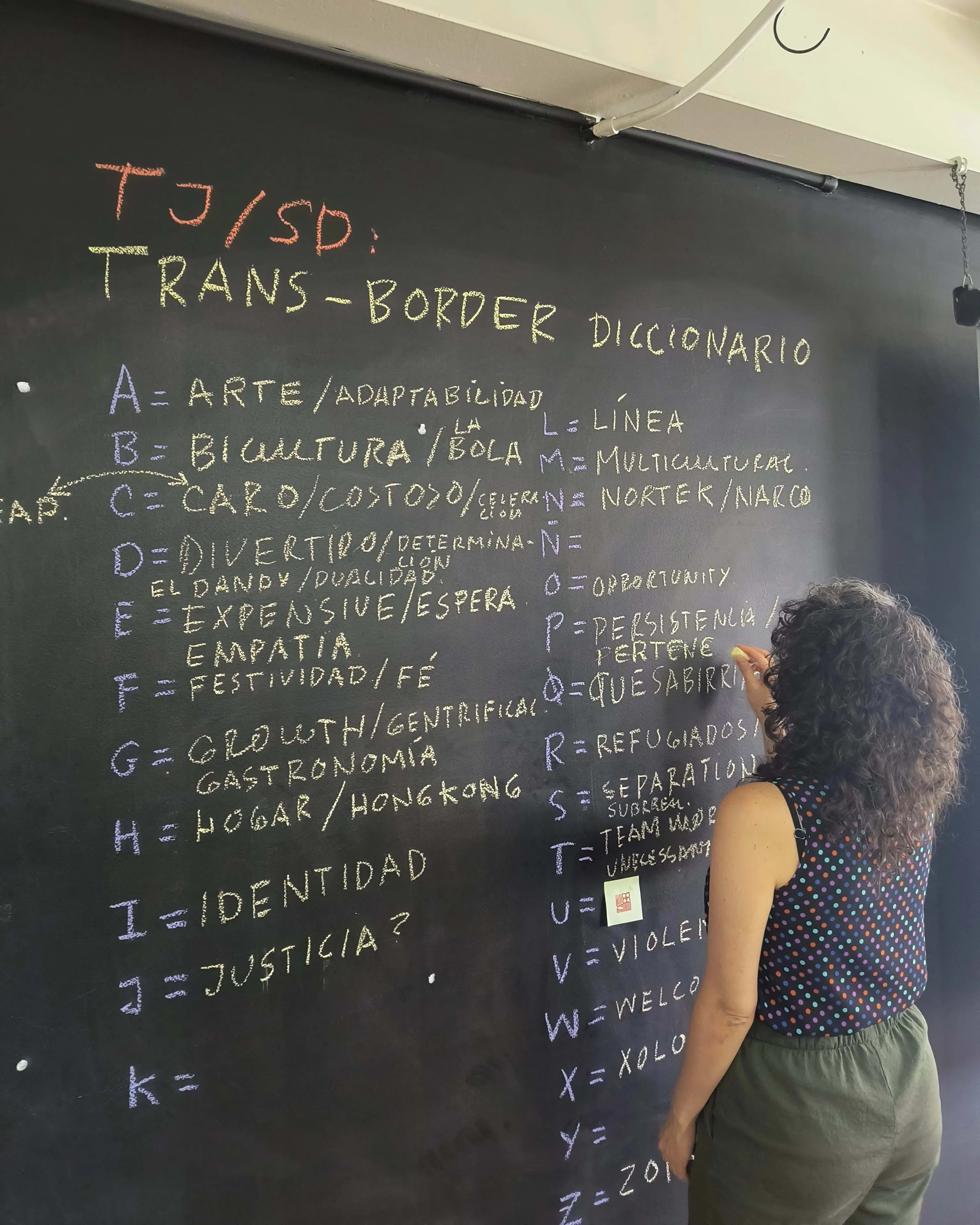 A woman writes Spanish words in yellow chalk on a blackboard next to letters of the alphabet written in purple chalk.