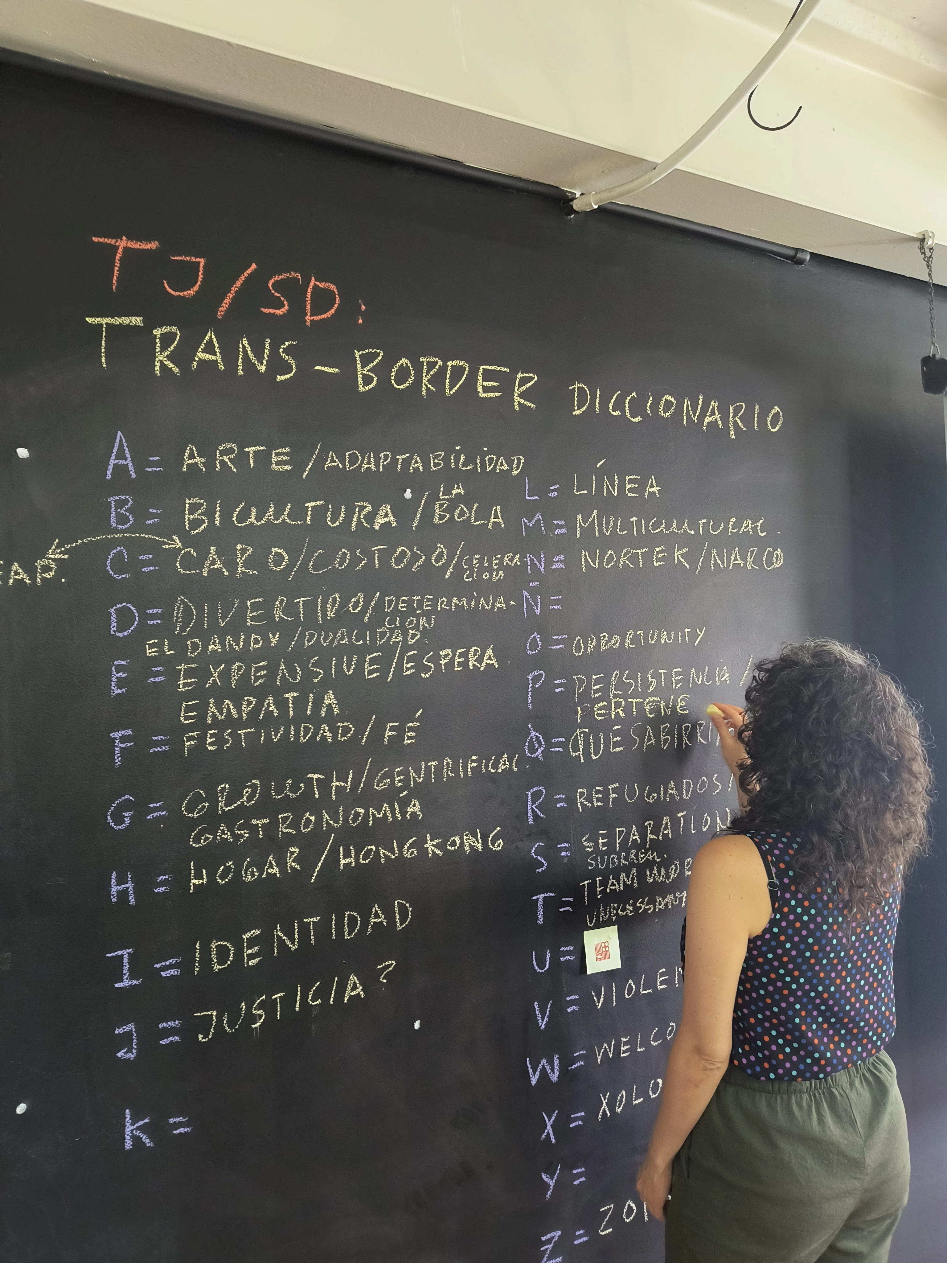 A woman writes Spanish words in yellow chalk on a blackboard next to letters of the alphabet written in purple chalk.