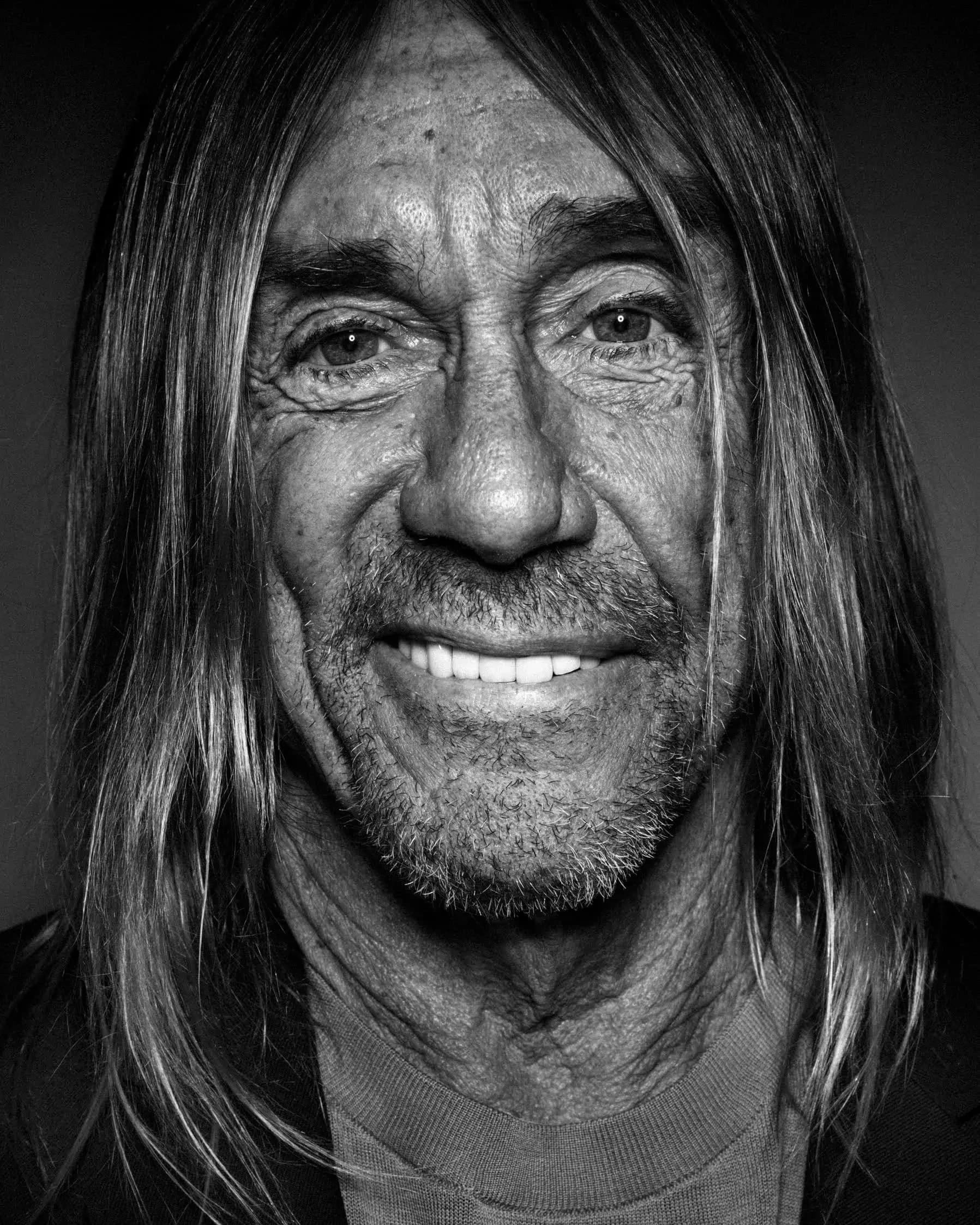 A black and white photo portrait of the singer Iggy Pop, who is looking straight into the camera with a smile. He has long hair and light skin, and is wearing a black cardigan over a gray t-shirt.