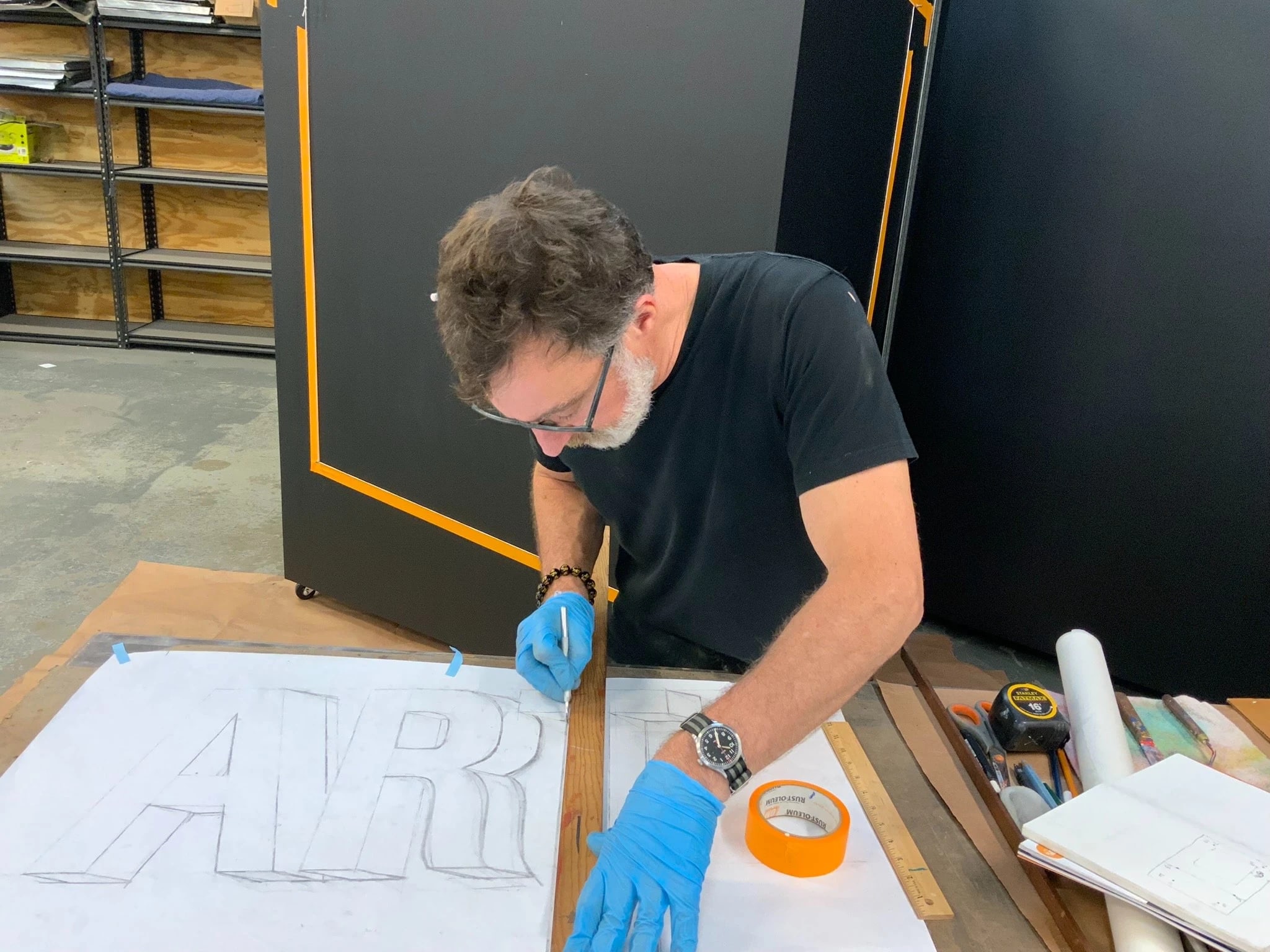 A man, stephen gaffney, working in his studio on a drawing that reads "AR.." on top of a wooden worktable. 