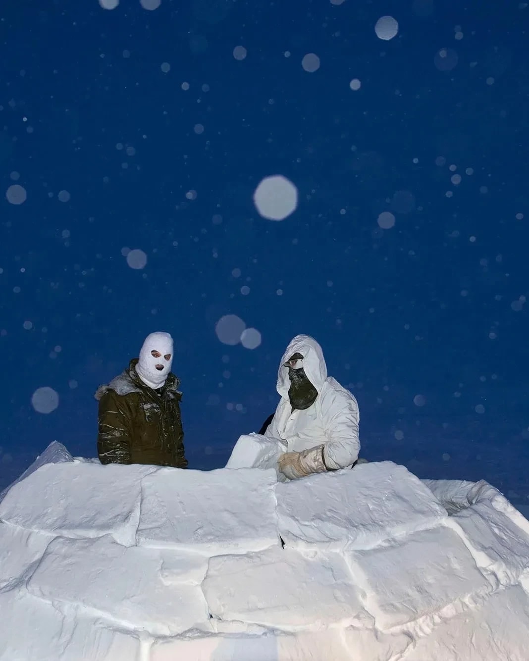 A color photograph of Two soldiers, wearing masks in an igloo made of snow blocks, at dusk while it snows, lit with flash.