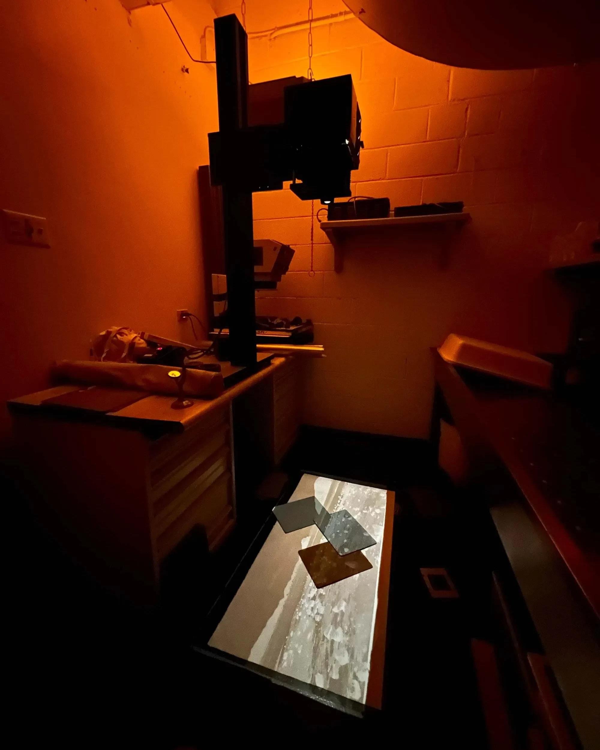 Dimly lit darkroom with an orange hue, featuring an enlarger projecting onto a surface. The emerging image creates an atmosphere of focus and mystery.