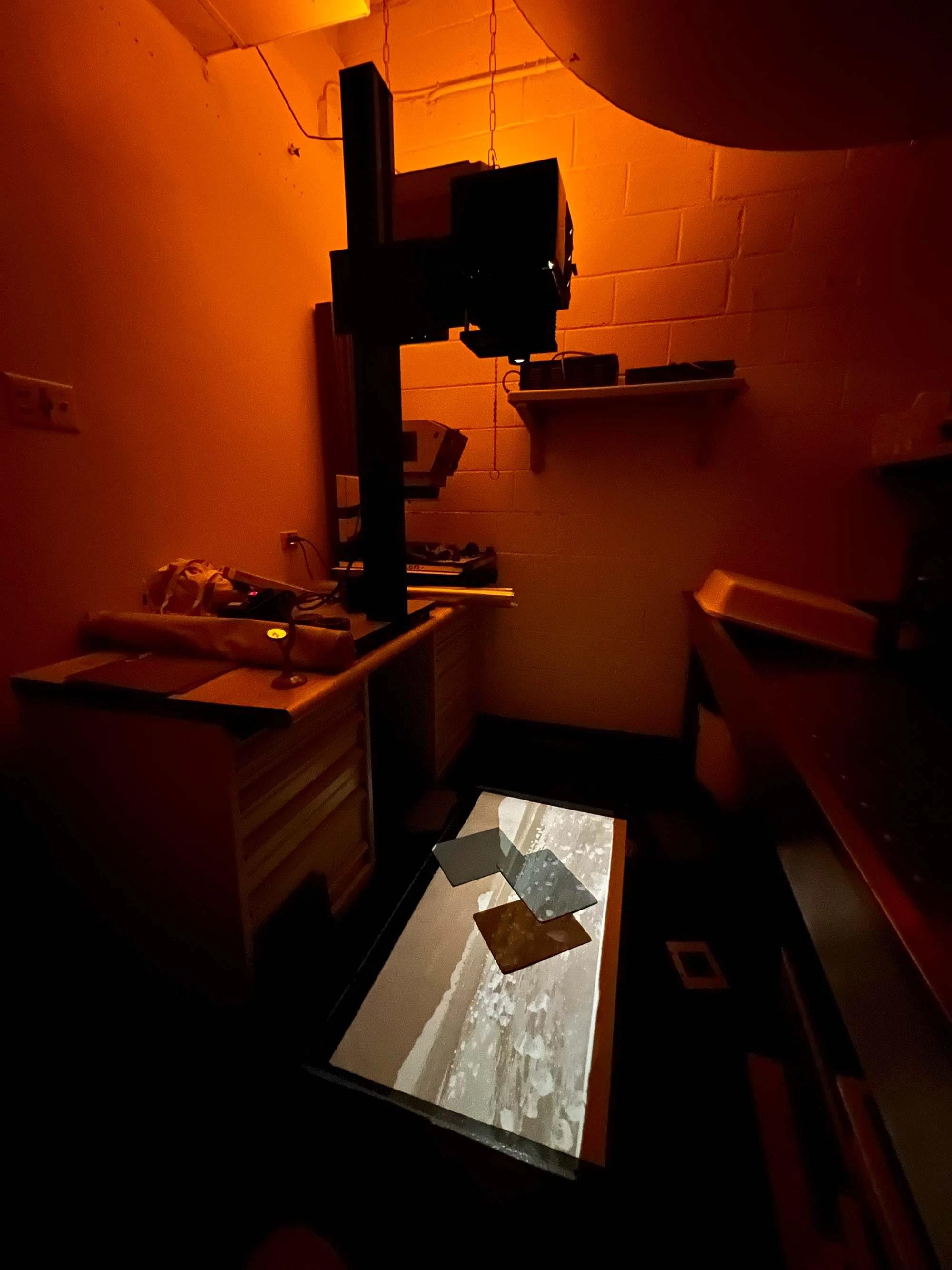 Dimly lit darkroom with an orange hue, featuring an enlarger projecting onto a surface. The emerging image creates an atmosphere of focus and mystery.