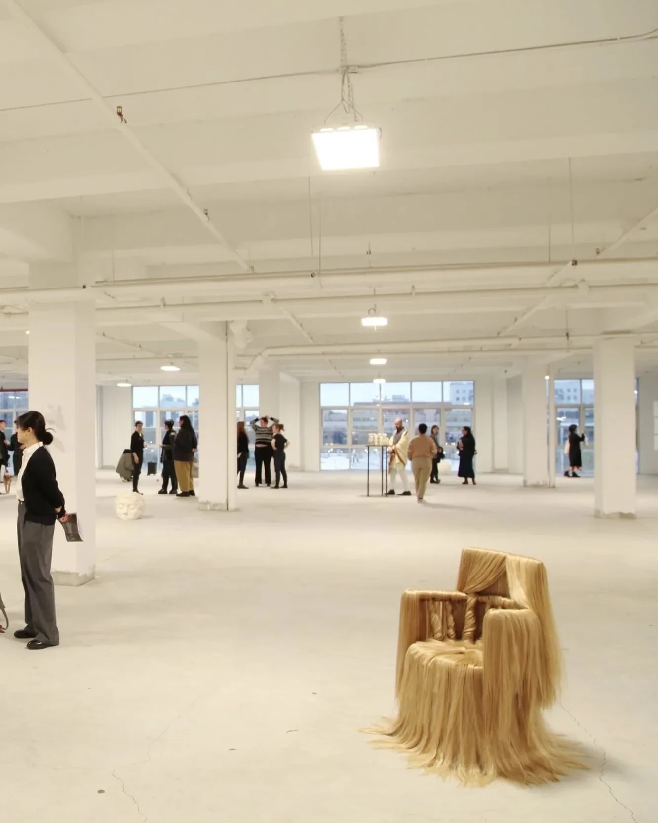 Wide view of an exhibition opening in a loft space with floor to ceiling windows. Many people mill about; there is a chair adorned with hair on display as well as art hanging on the wall.