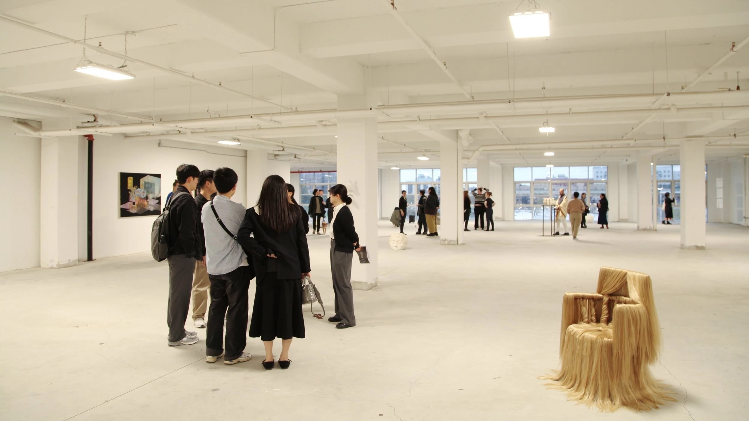 Wide view of an exhibition opening in a loft space with floor to ceiling windows. Many people mill about; there is a chair adorned with hair on display as well as art hanging on the wall.