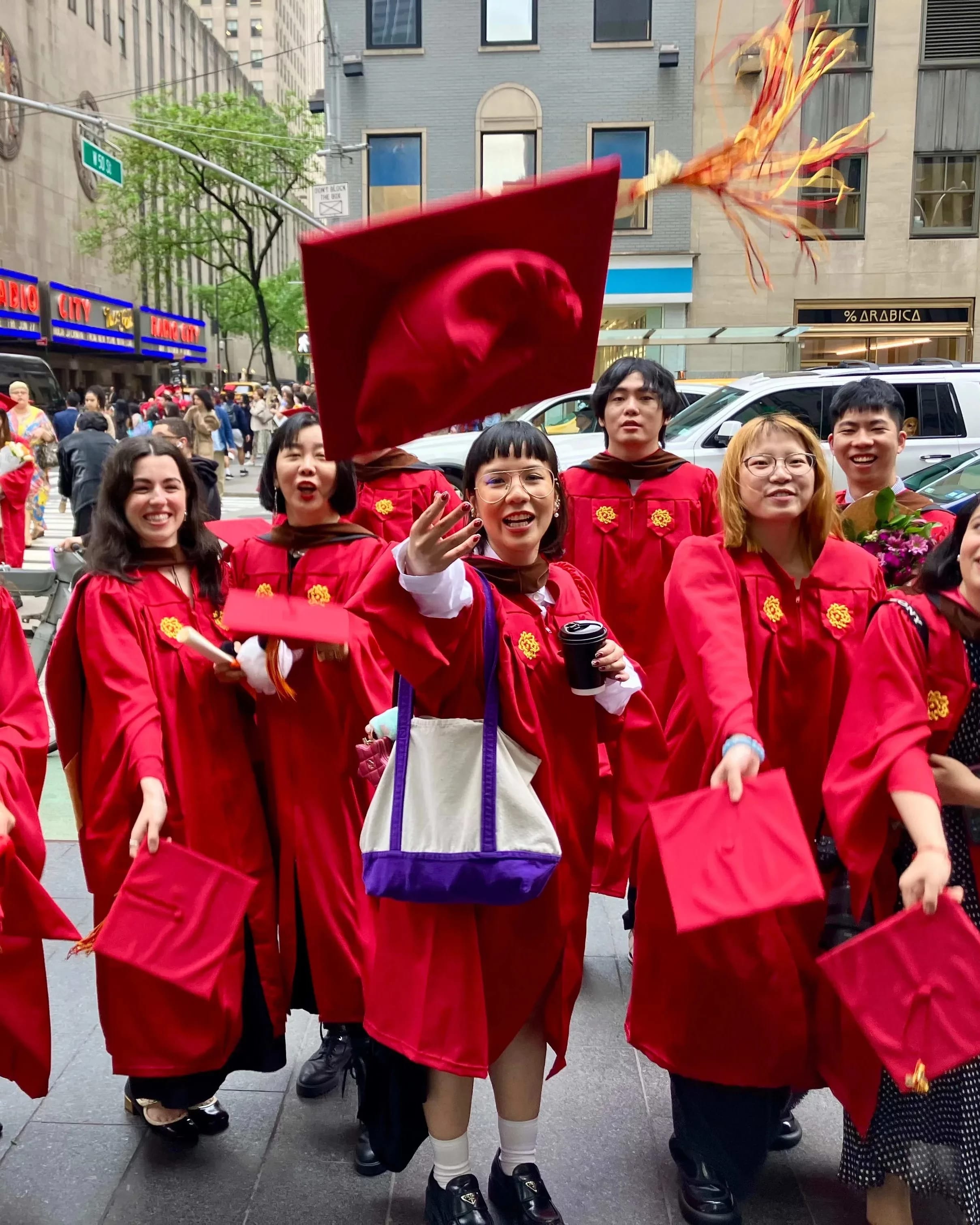 The Masters in Digital Photography graduating class of 2025, in the iconic SVA red graduation gowns, in front of the famed Radio City Music Hall where the commencement ceremony just took place. The students are threwing their caps in excitement. 