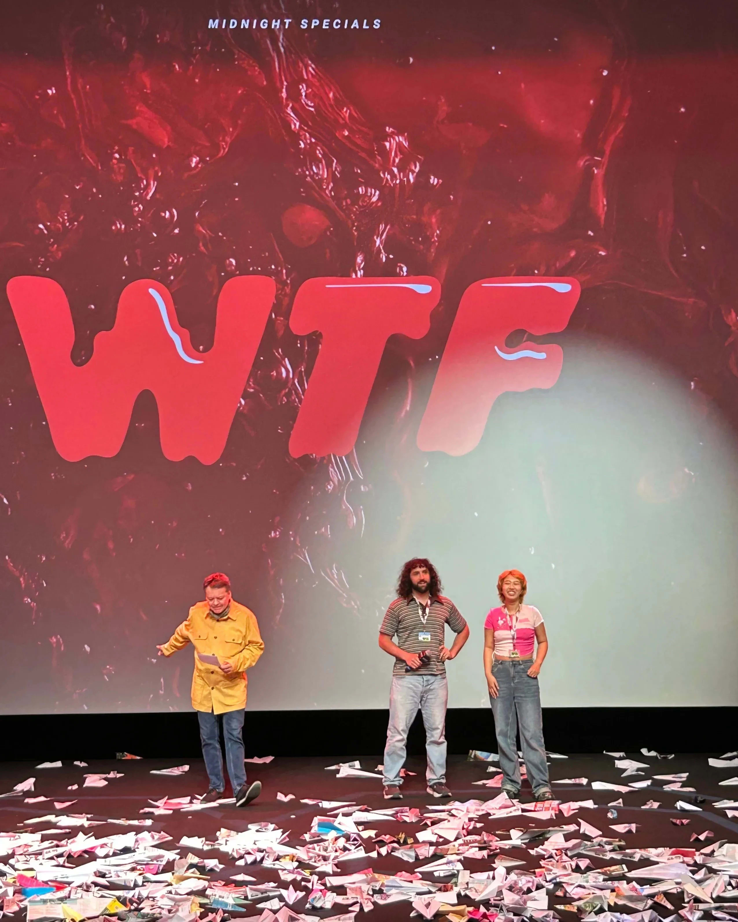 Three people stand on a stage full of paper airplanes with a projection showing WTF behind them