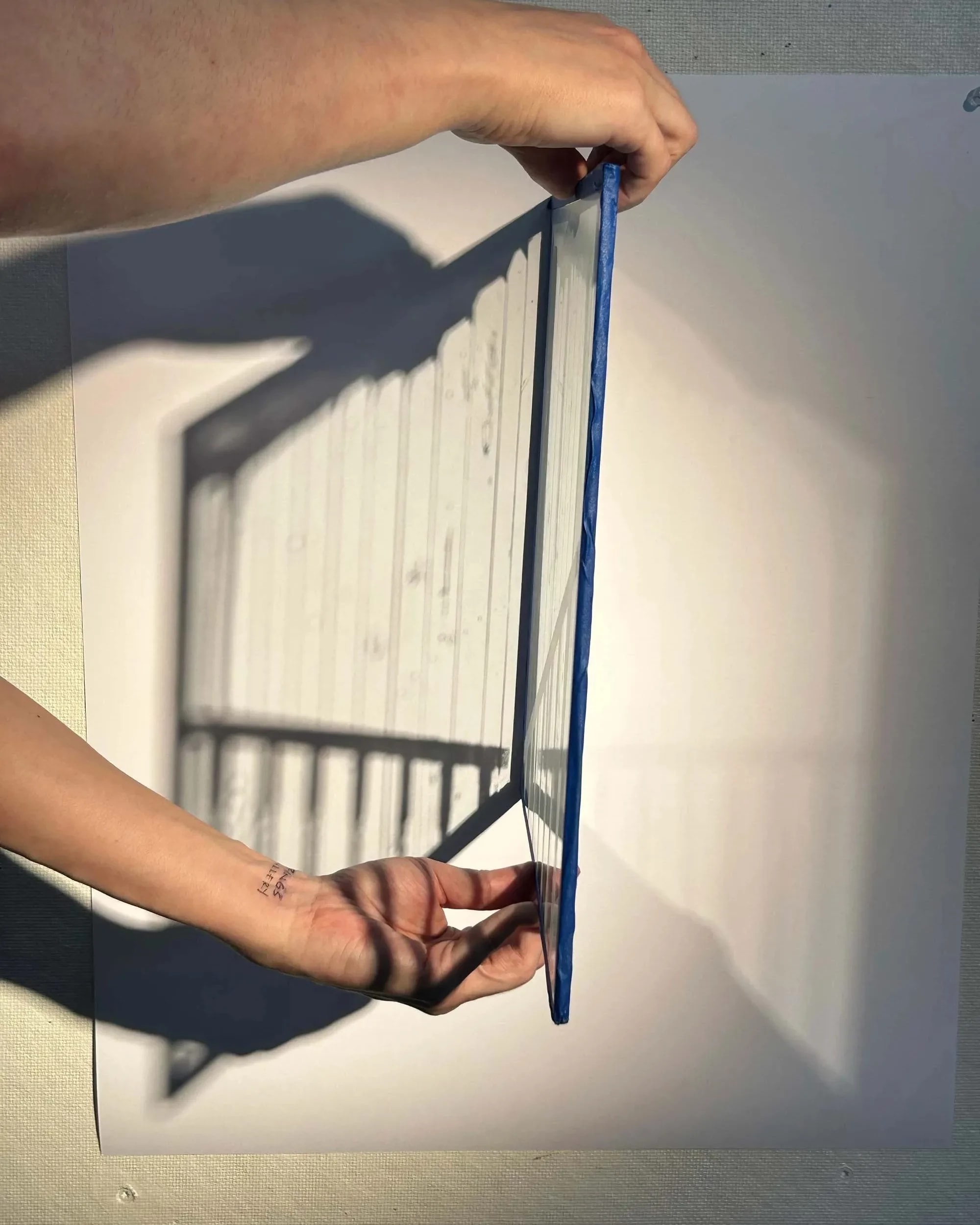 Hands hold a transparent, blue-edged panel casting a lined shadow on a white surface. The scene is minimalistic and abstract.