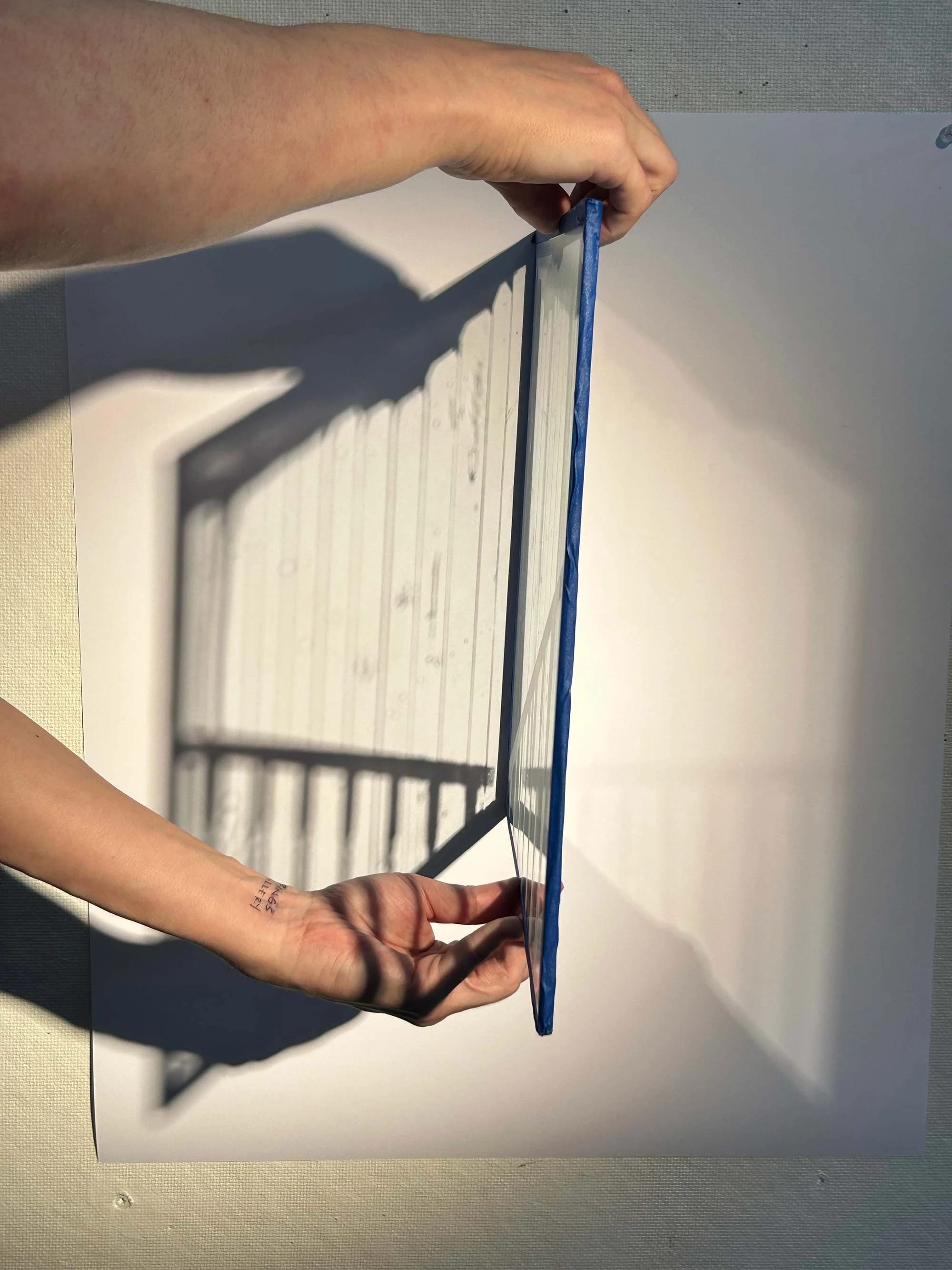 Hands hold a transparent, blue-edged panel casting a lined shadow on a white surface. The scene is minimalistic and abstract.