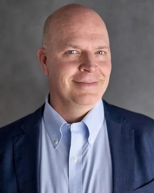 A man in a sky blue collared shirt and darker blue suit jacket smiles into 