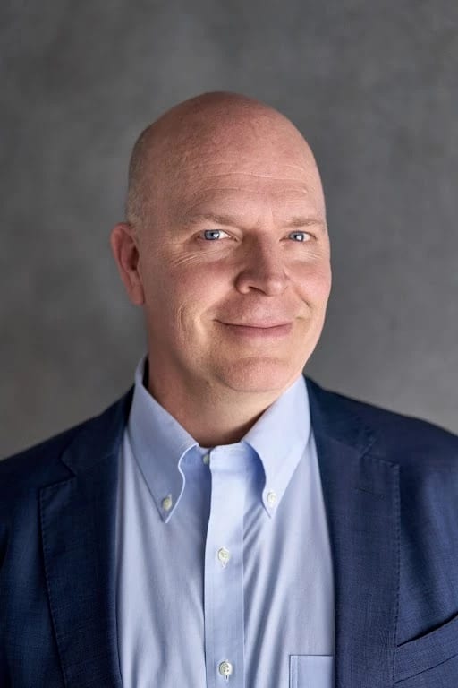 A man in a sky blue collared shirt and darker blue suit jacket smiles into 