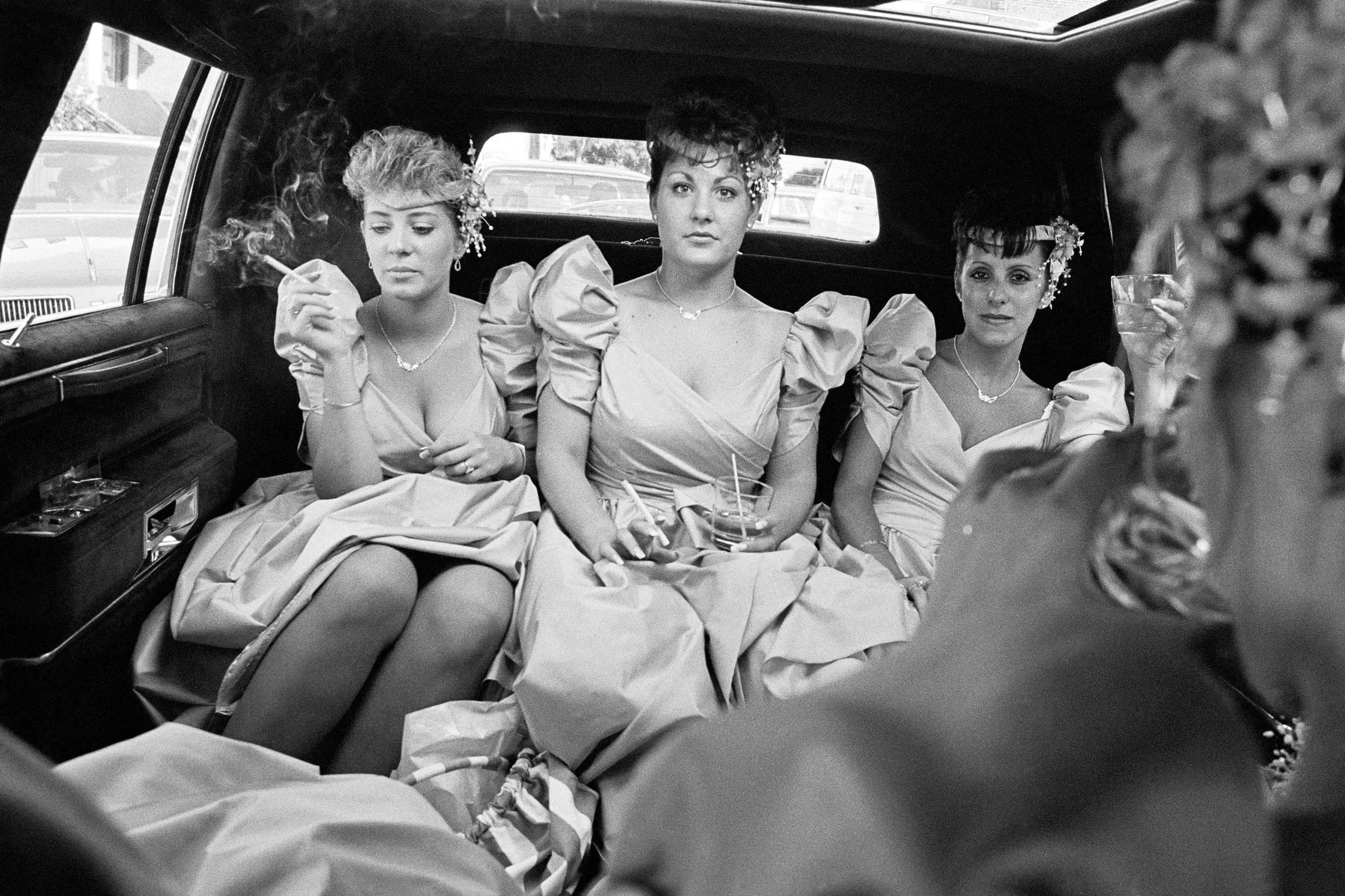 Three women in elegant dresses seated in the back of a car, enjoying a moment together during their journey.