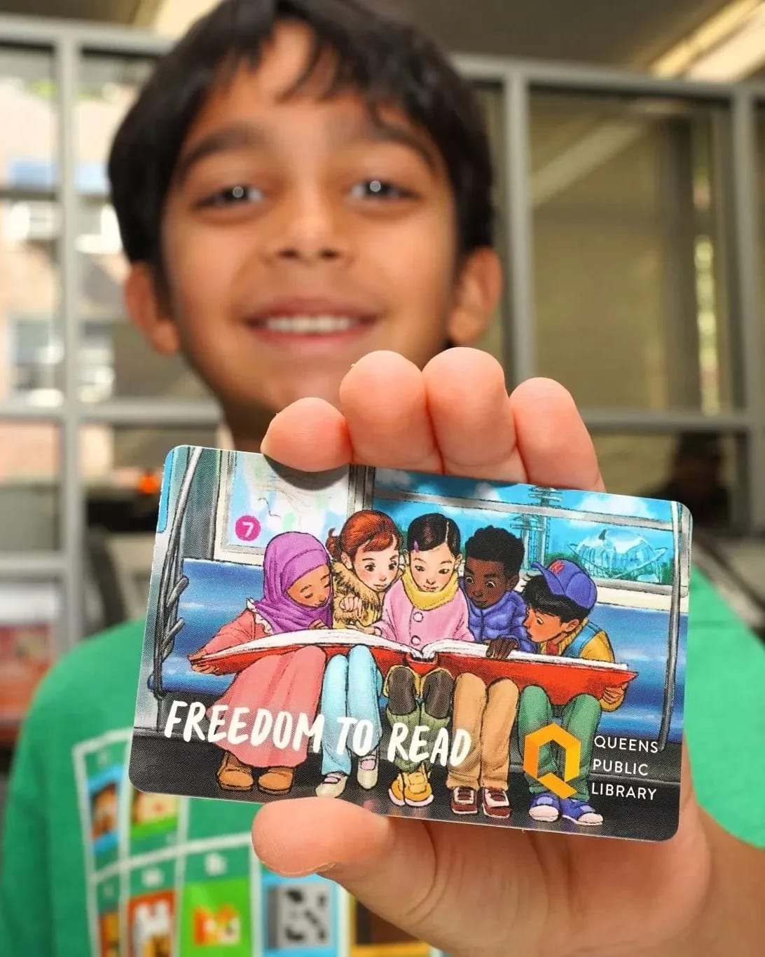 A little boy smiles as he holds up his new library card that has the illustration by Sammi Wu.