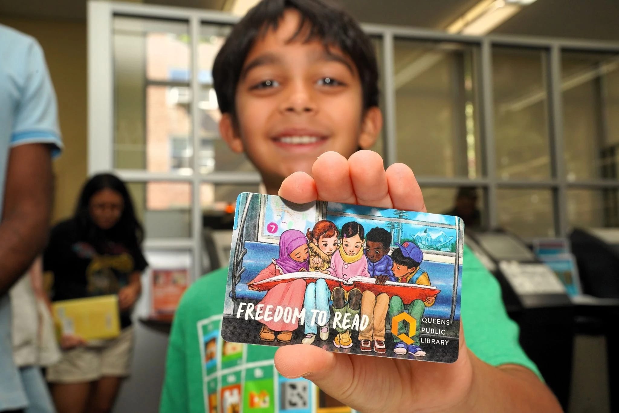 A little boy smiles as he holds up his new library card that has the illustration by Sammi Wu.