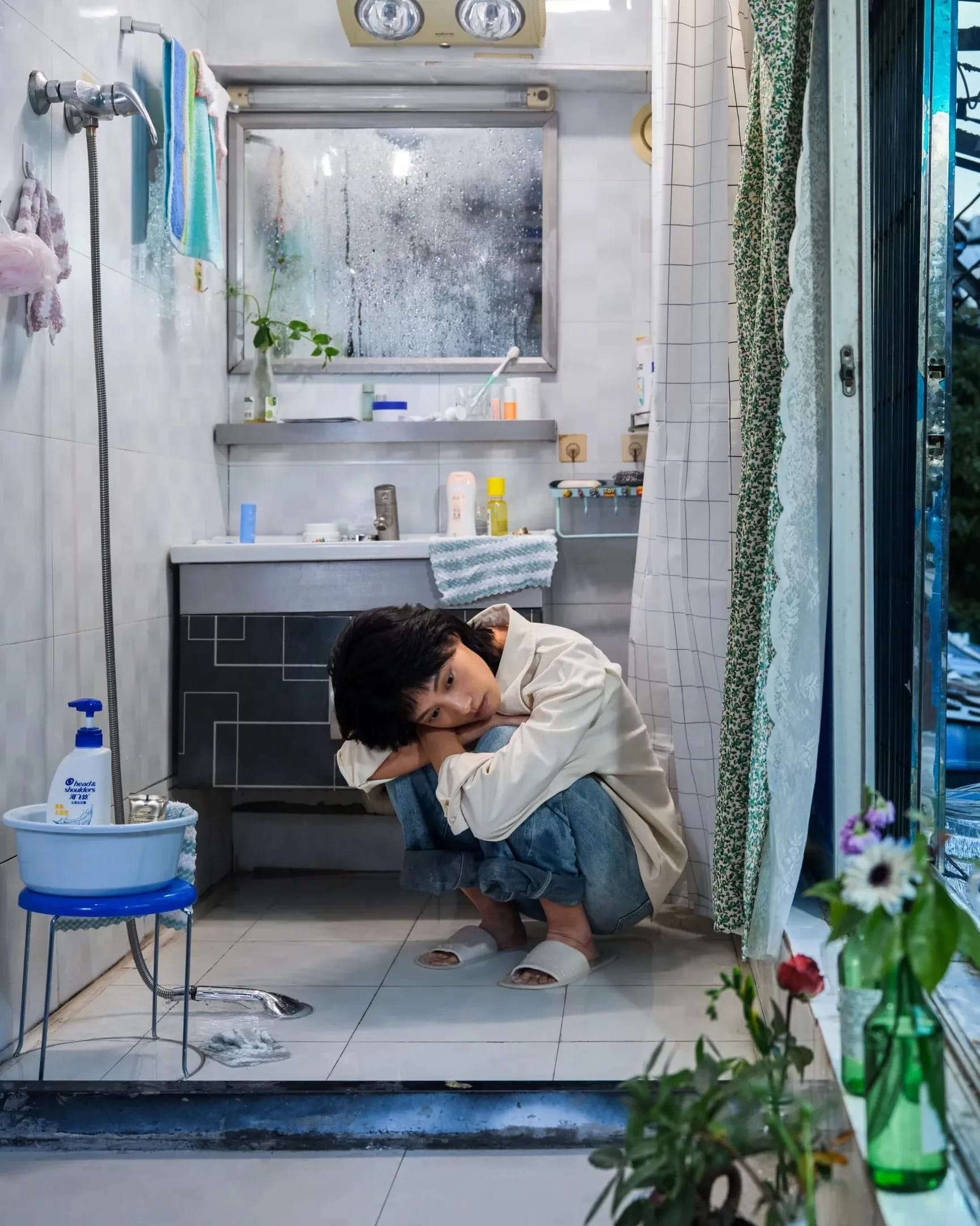 A color photography by Shaohan Fang entitled En Route. In a dim bathroom at dusk, the girl crouches on a wet floor, staring at the window. Water pools around her feet, she appears visibly overwhelmed.