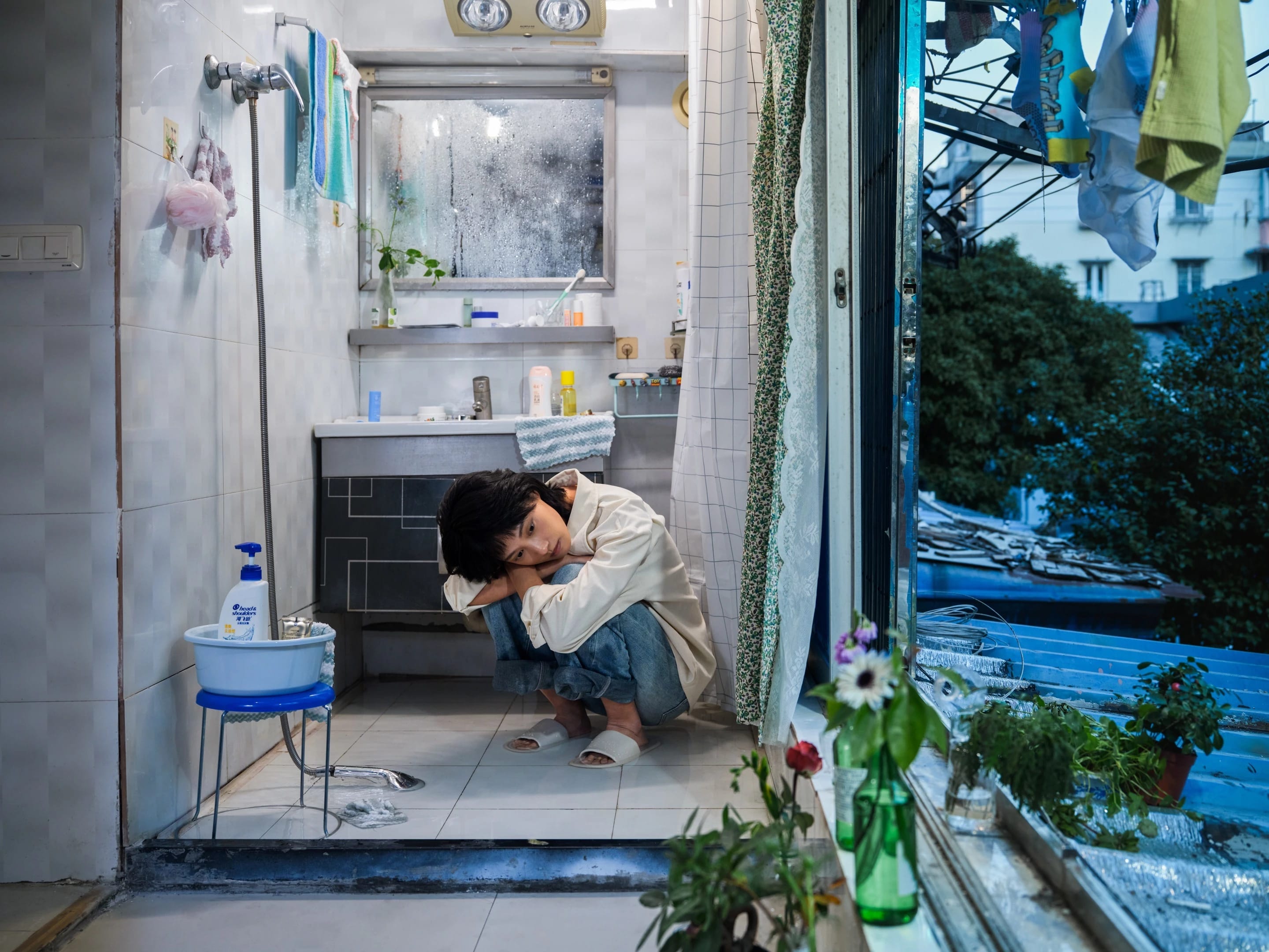 A color photography by Shaohan Fang entitled En Route. In a dim bathroom at dusk, the girl crouches on a wet floor, staring at the window. Water pools around her feet, she appears visibly overwhelmed.