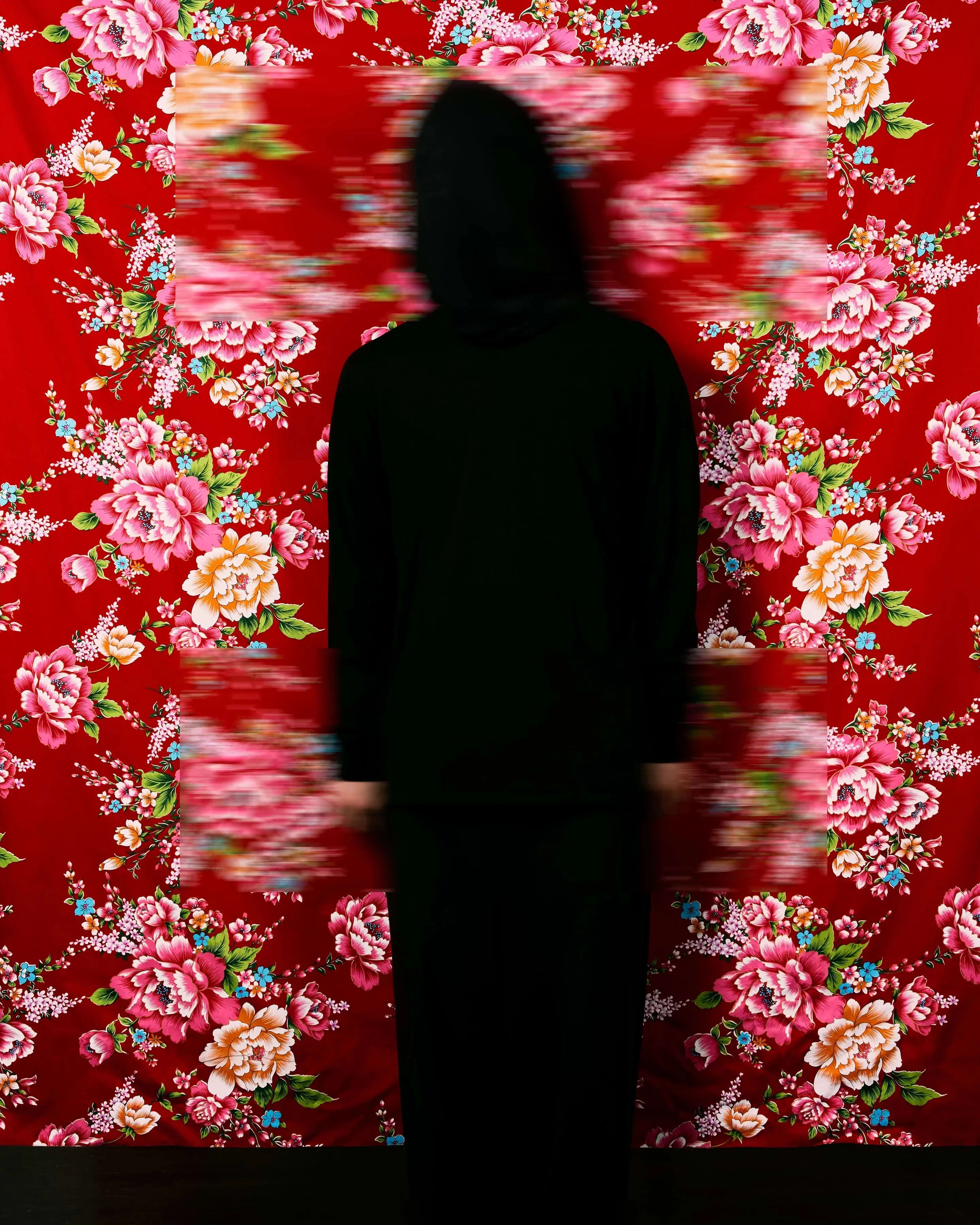 A color photograph featuring a figure wrapped in black from head to toe standing facing the camera against a red Hakka floral fabric background, which is suspended and reveals a faint dark wooden floor below. Three blurred rectangles cover the head, hands, and feet.