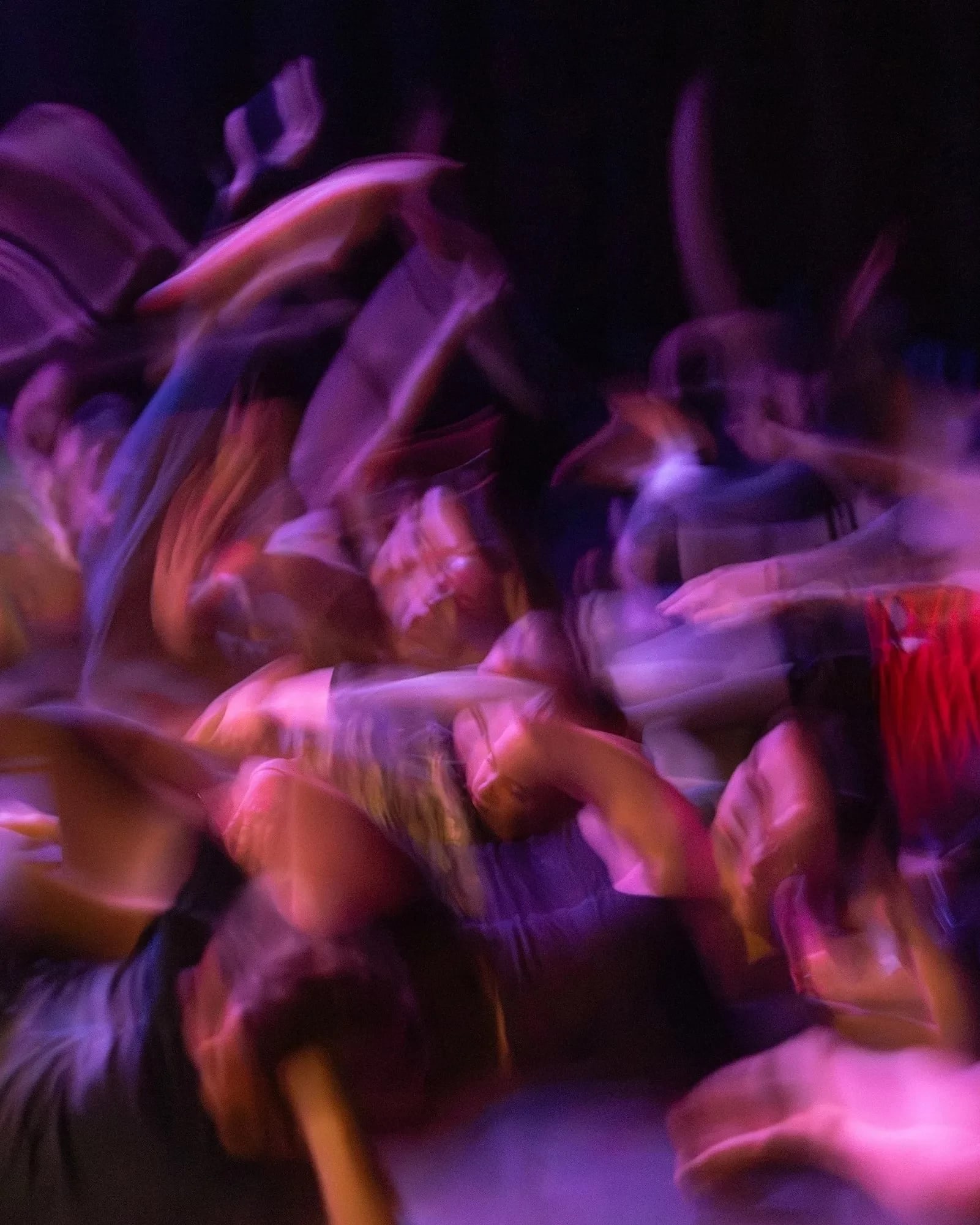 A dynamic, motion-blurred photograph of a crowd in movement under purple and amber stage lighting, suggesting dance or performance.