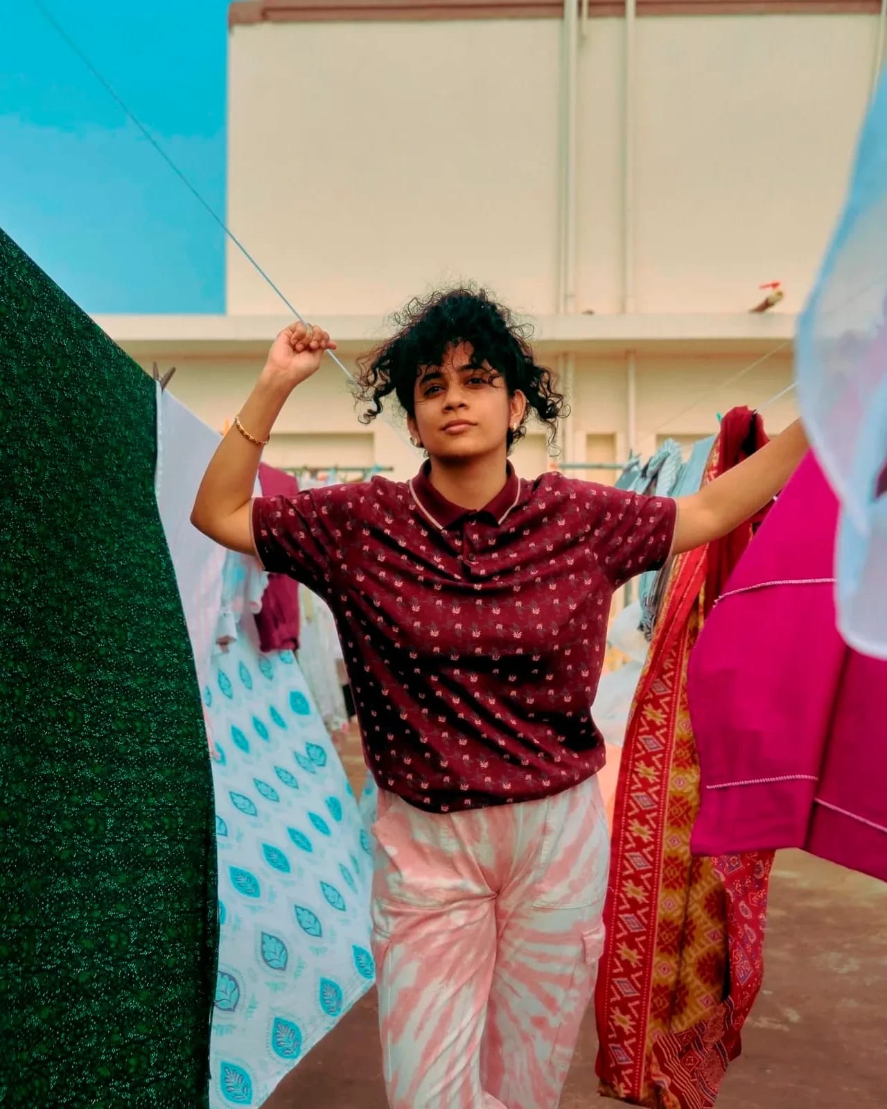 In this color self-portrait, the artist Shravya Kag is standing among pieces of fabric drying on cloth lines. She is wearing a short sleeve top in burgundy red with yellow polka dots, and a pair of tie-dyed long pants in salmon pink and white.