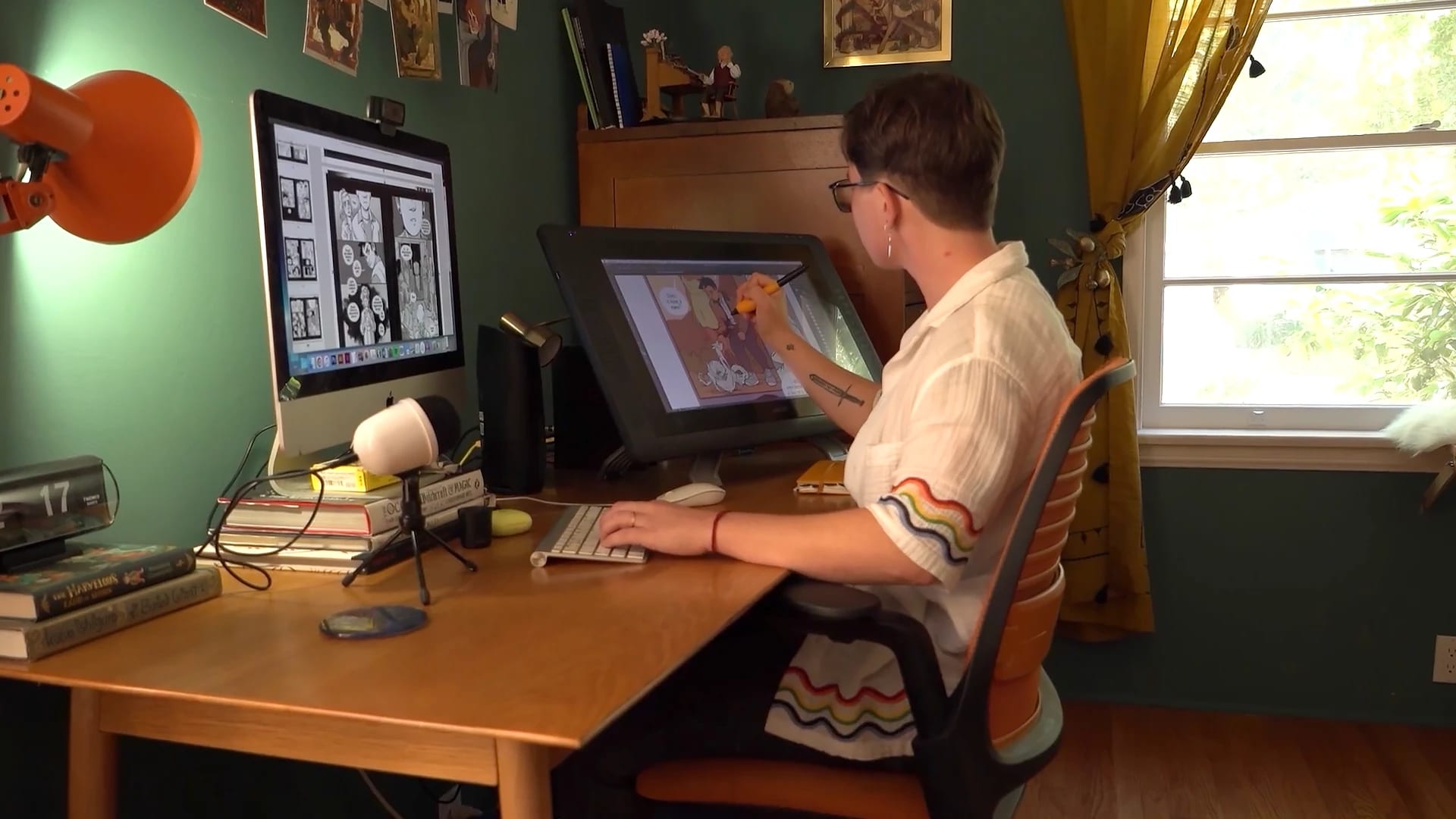 Molly Knox Ostertag sits in front of a Mac and a Wacom Cintiq. The walls of the room are green and there is a window behind them.