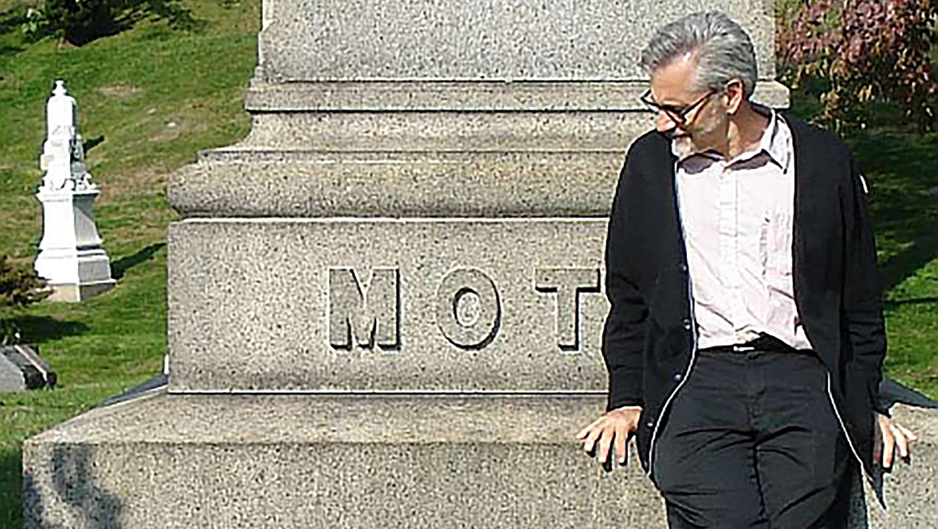 Cartoonist tom motley in a graveyard sitting on a large memorial gravestone that says "MOT"