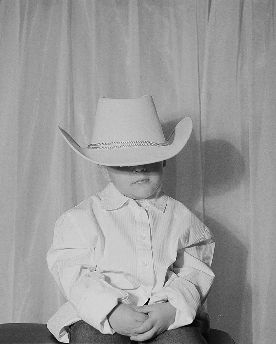 Black and white photo of a child wearing a cowboy hat that is too big for them, obscuring their face.