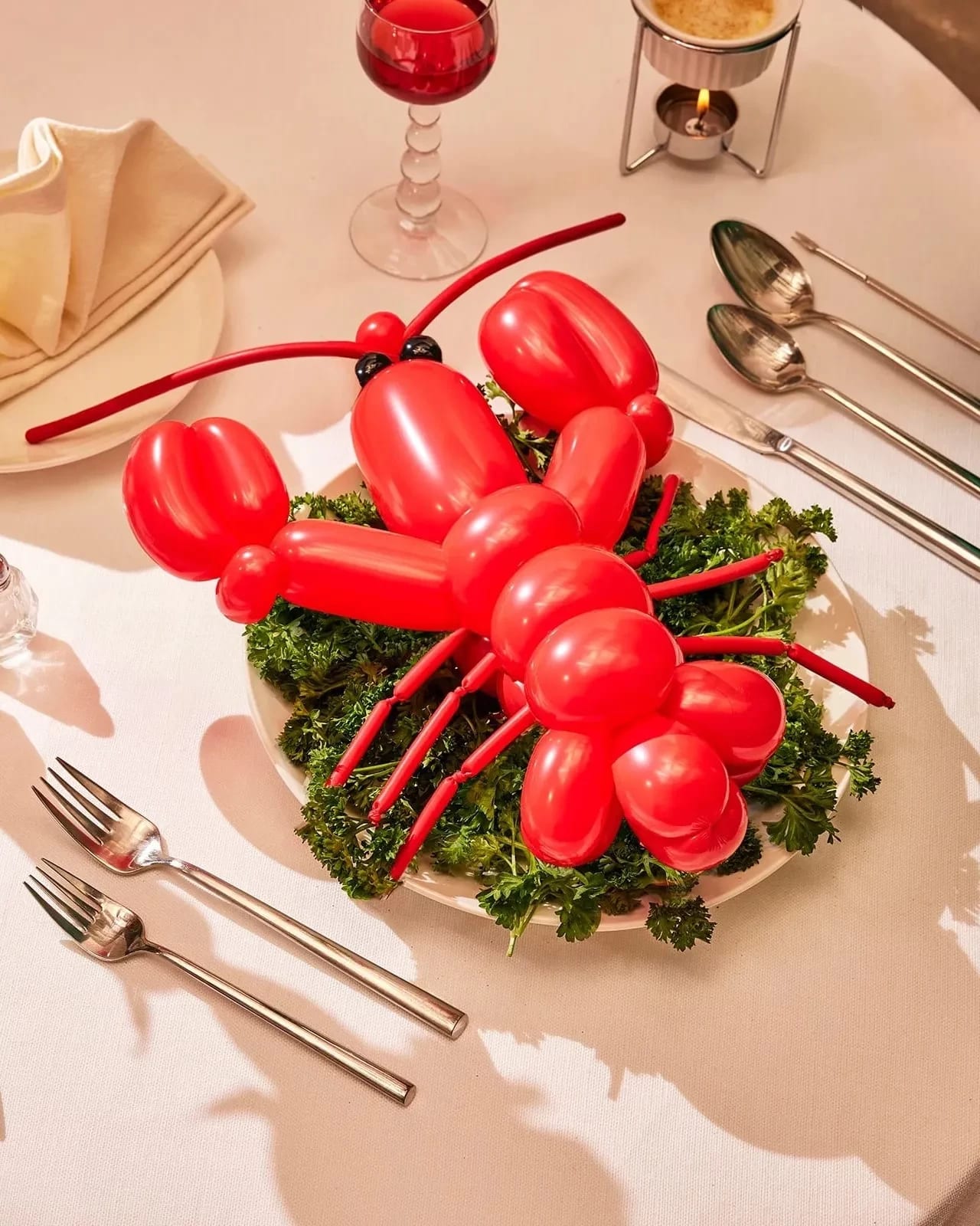 A photograph of a lobster shaped red balloon sitting on top of a bed of kale leaves on a dinner plate. Surrounding the scene is a dinner table fully set up with cutlery,  a dinner napkin folded into a fan, and a single rose in a tin vase.