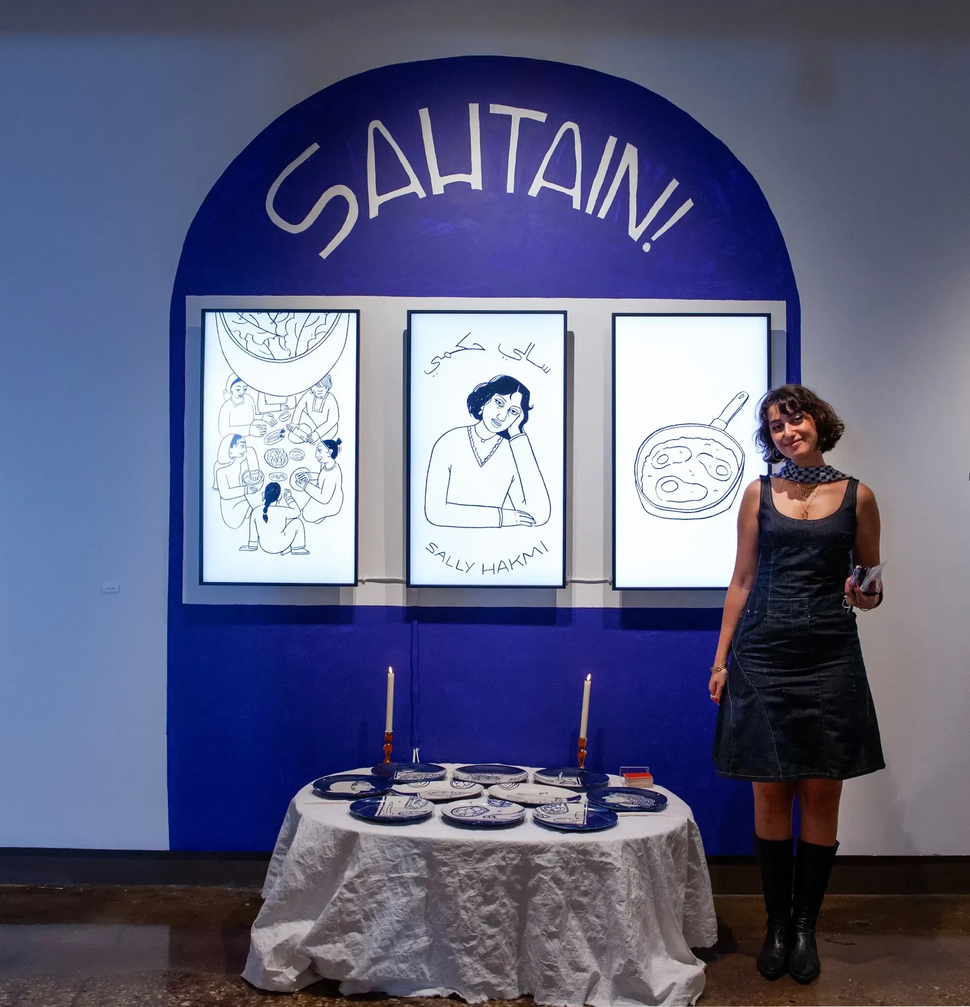 A person stands in front of an exhibit featuring a table dressed with plates and candles in front of a wall with a painted blue arch and three screens showing off illustrations.