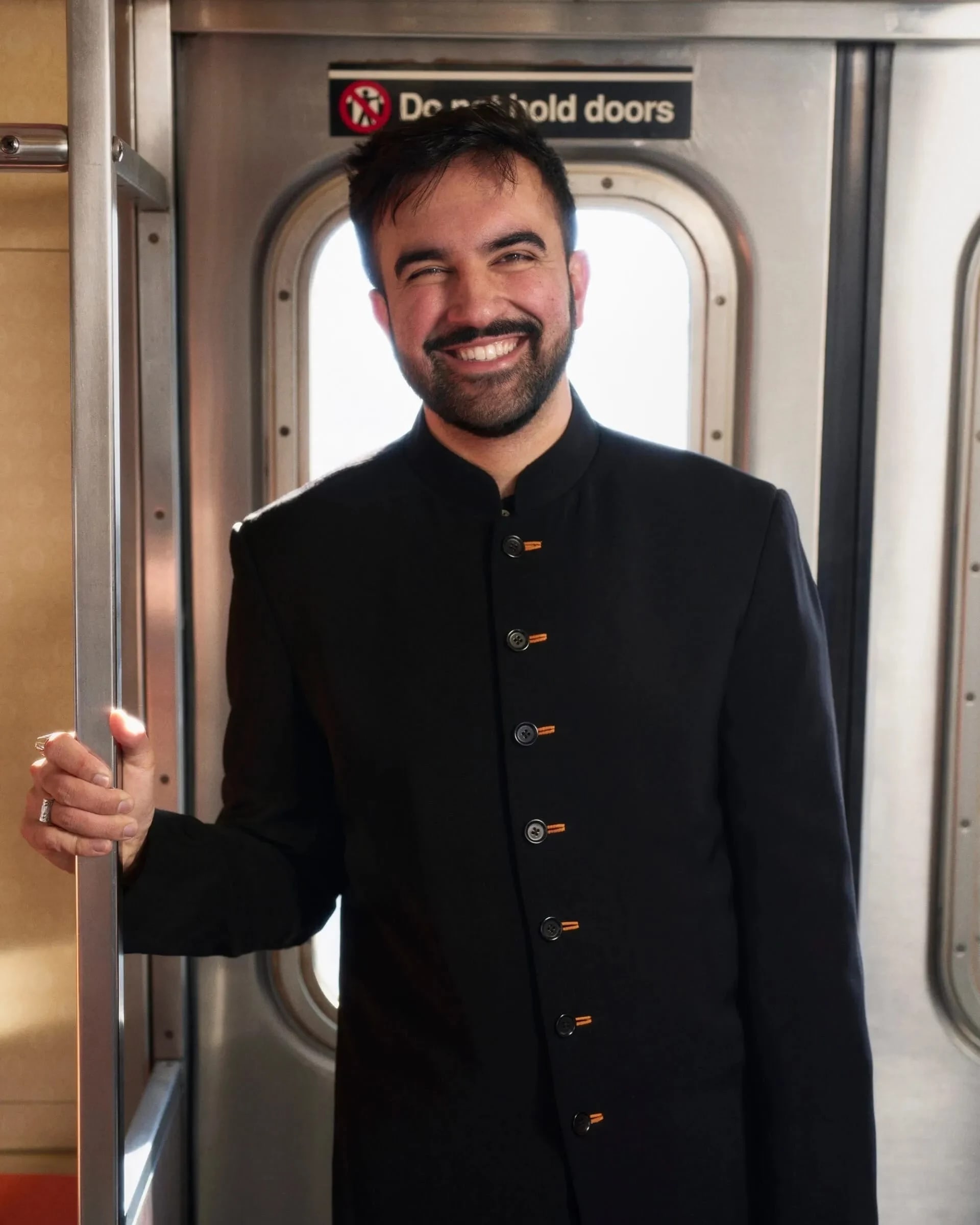 A person stands on the subway in a black button up. They are smiling widely and warmly.