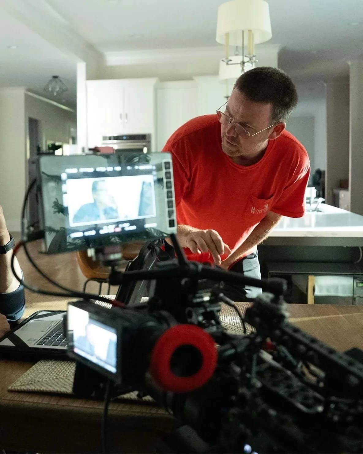 A man seated at a table in a modern kitchen, focused on a laptop, while another man in a red shirt leans over, discussing something, with camera equipment in the foreground.