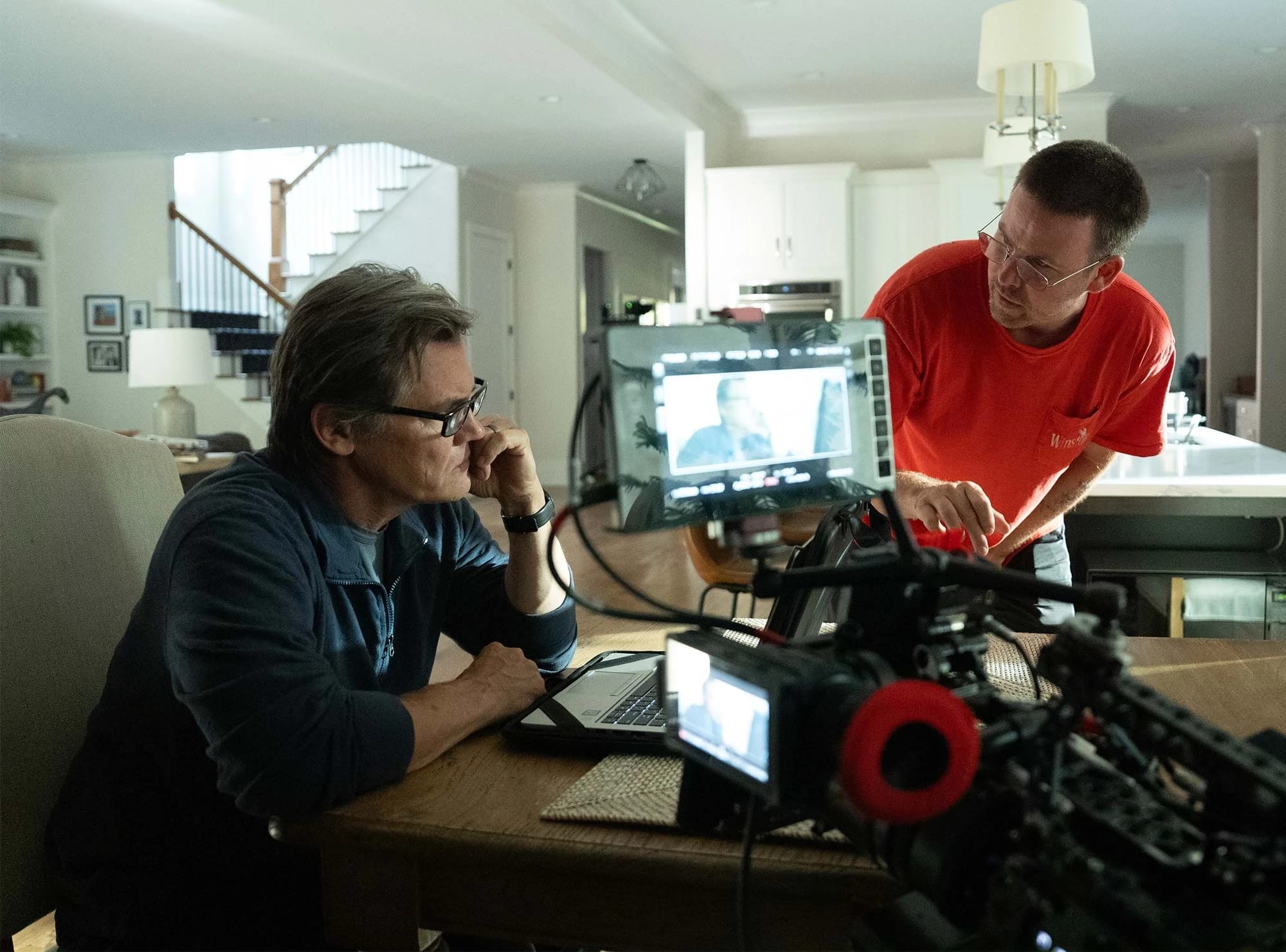 A man seated at a table in a modern kitchen, focused on a laptop, while another man in a red shirt leans over, discussing something, with camera equipment in the foreground.