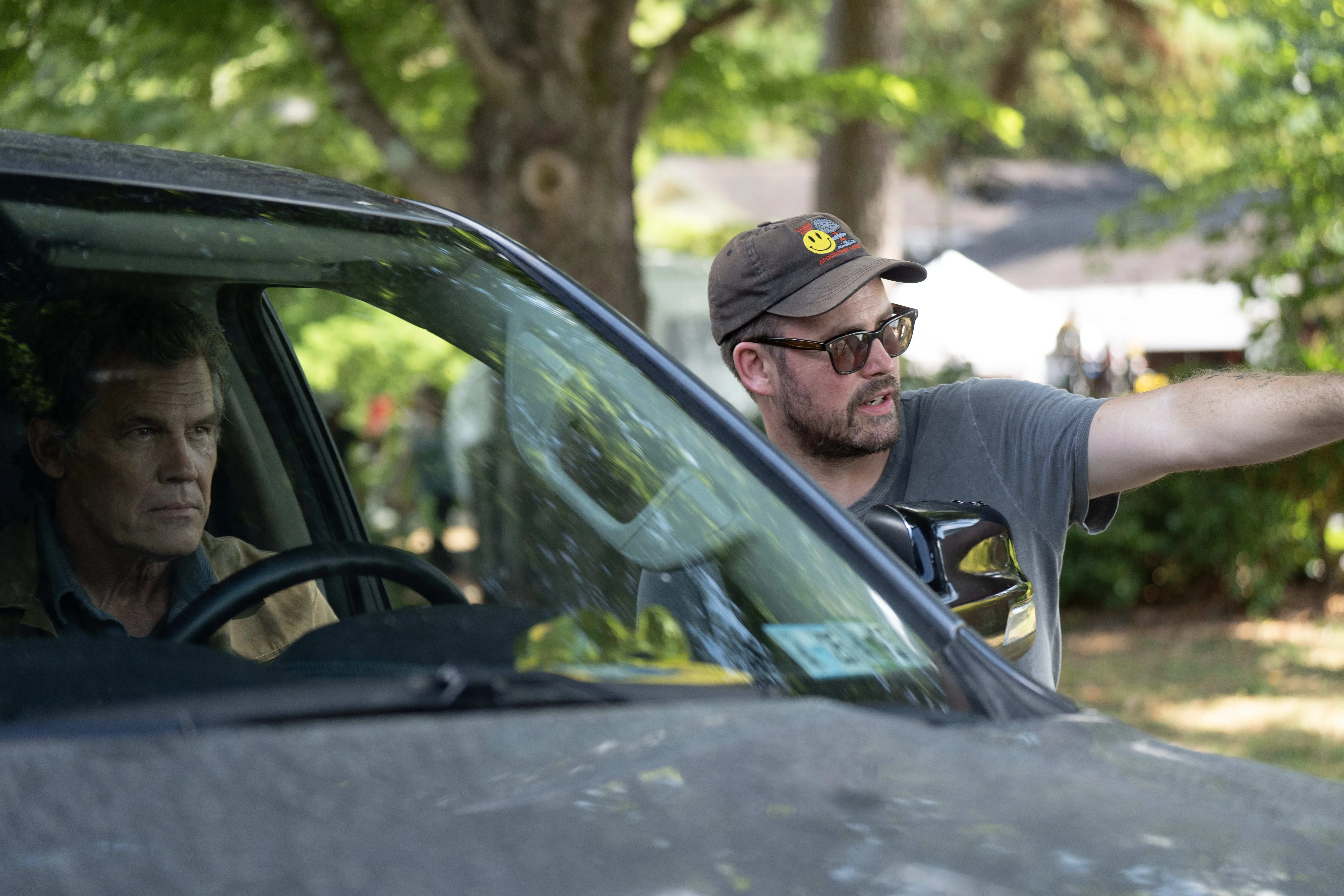 A man sits focused in a car, looking forward. Another man outside gestures and directs, wearing glasses and a cap. Trees create a serene, green backdrop.
