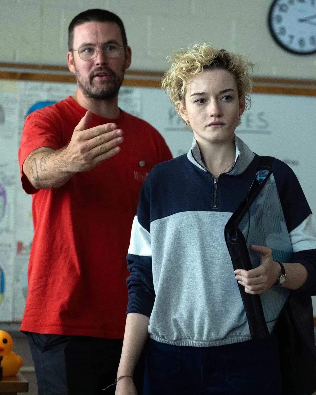 A person in a red shirt gestures in a classroom, appearing explanatory, while another with short curly hair listens intently. A whiteboard and clock are in the background.