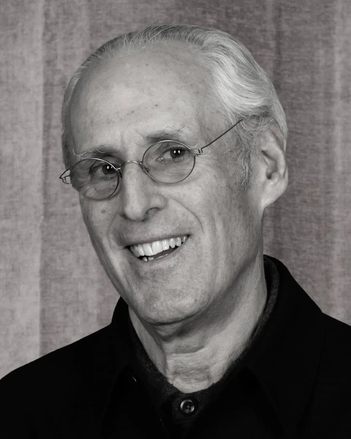 A person with short white hair and glasses smiles at the camera. He is wearing a dark jacket in front of a plain, textured background.