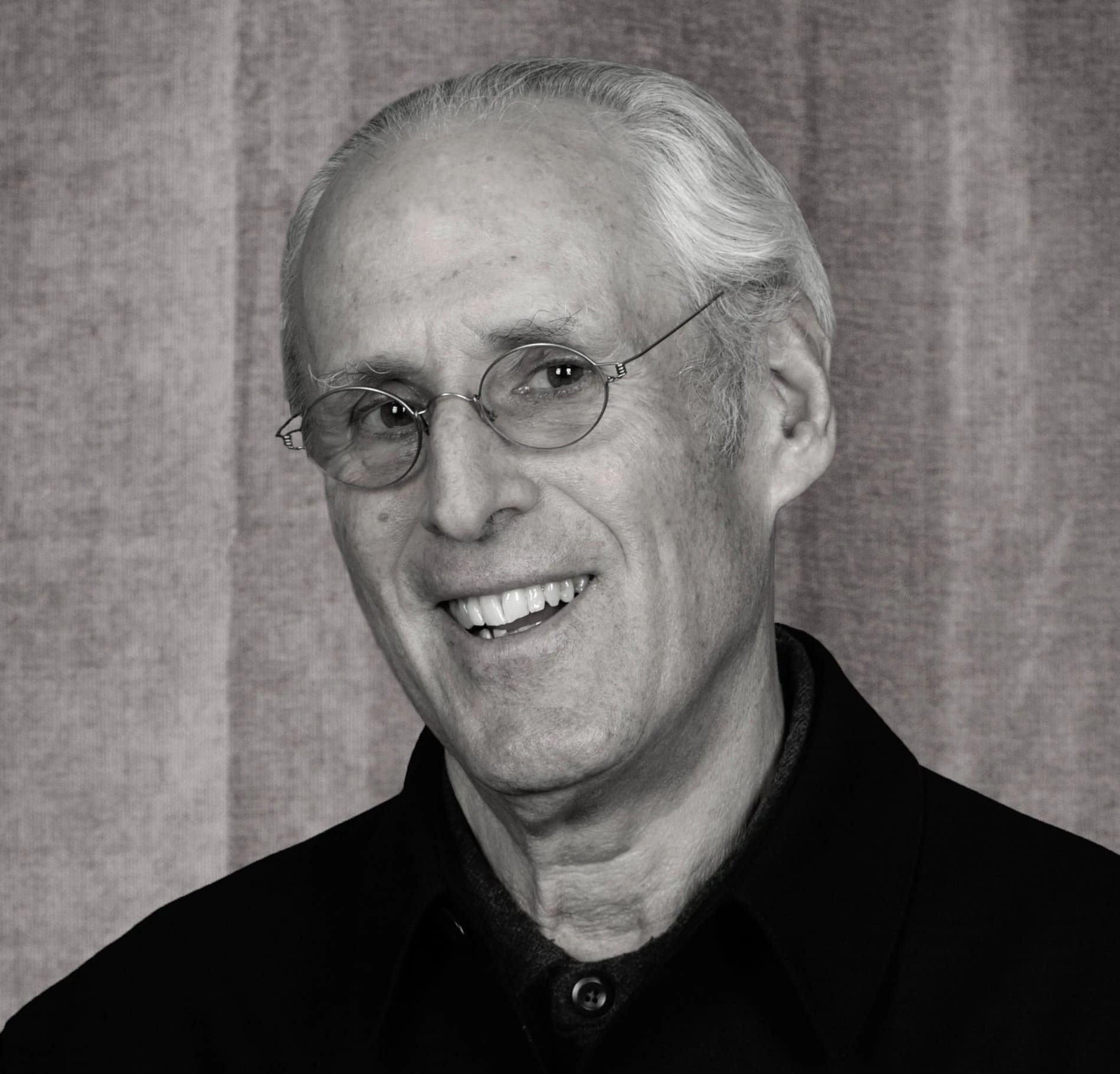A person with short white hair and glasses smiles at the camera. He is wearing a dark jacket in front of a plain, textured background.