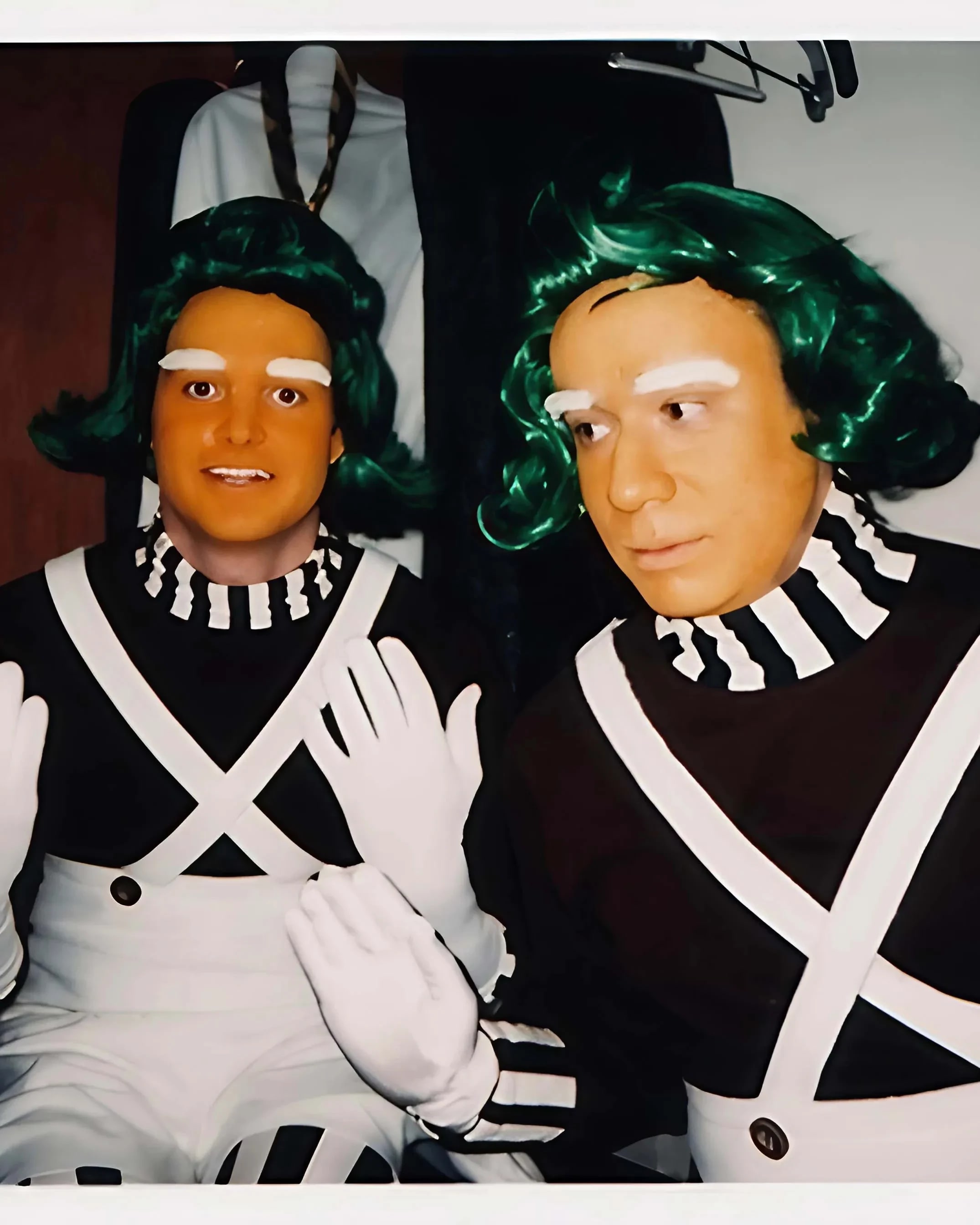 Two men dressed in elaborate costumes, sporting green hair and white gloves, display a cheerful demeanor in the image.