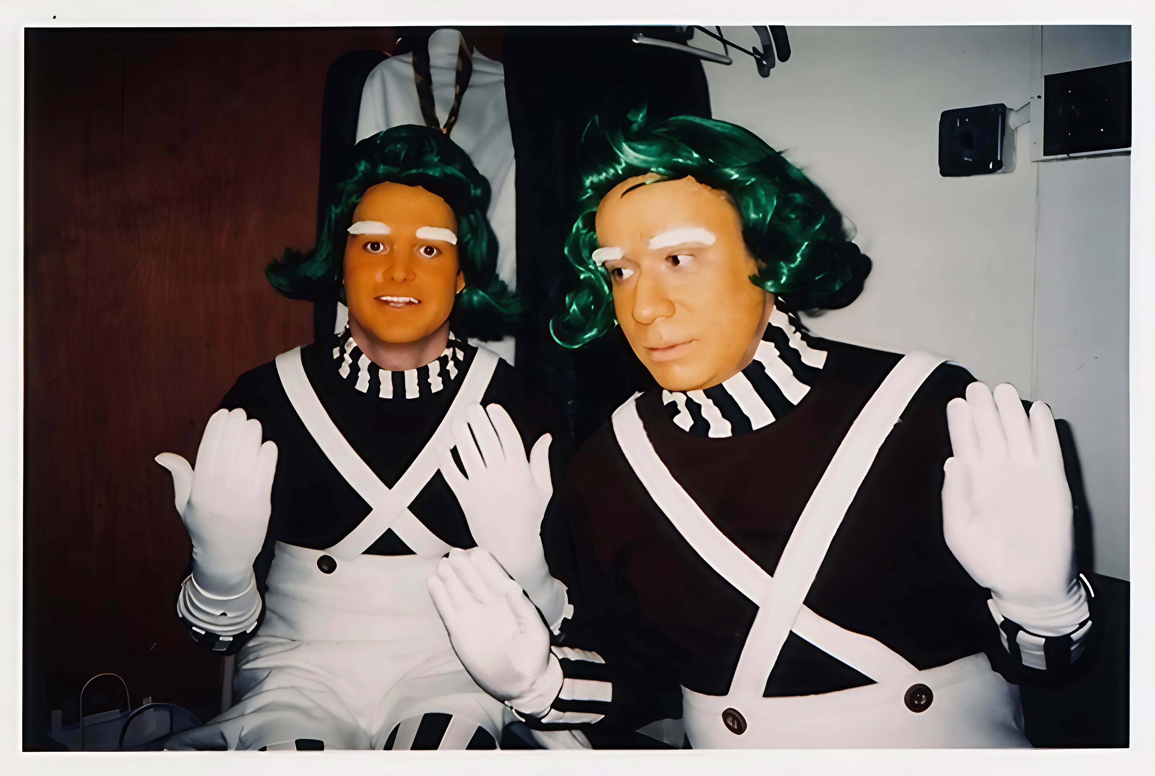 Two men dressed in elaborate costumes, sporting green hair and white gloves, display a cheerful demeanor in the image.