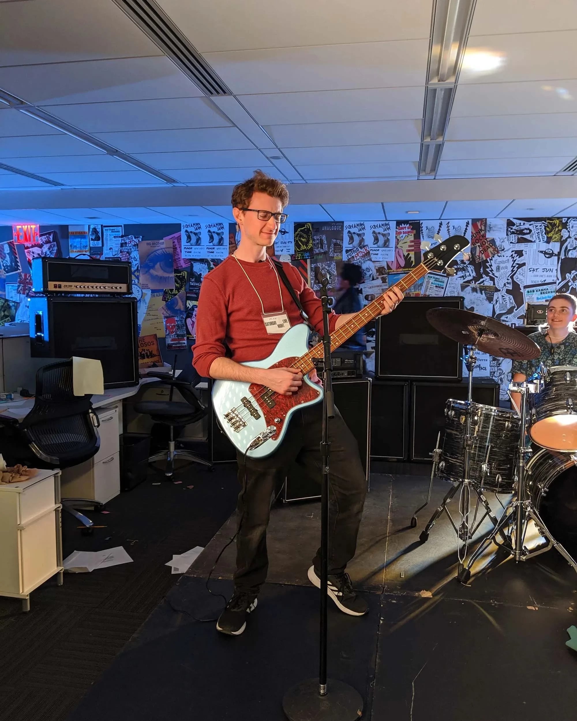A musician playing a blue bass guitar while standing on stage. Behind him, a drummer is seated, and the background features posters on the walls. The setting appears to be a music space with various instruments and equipment.