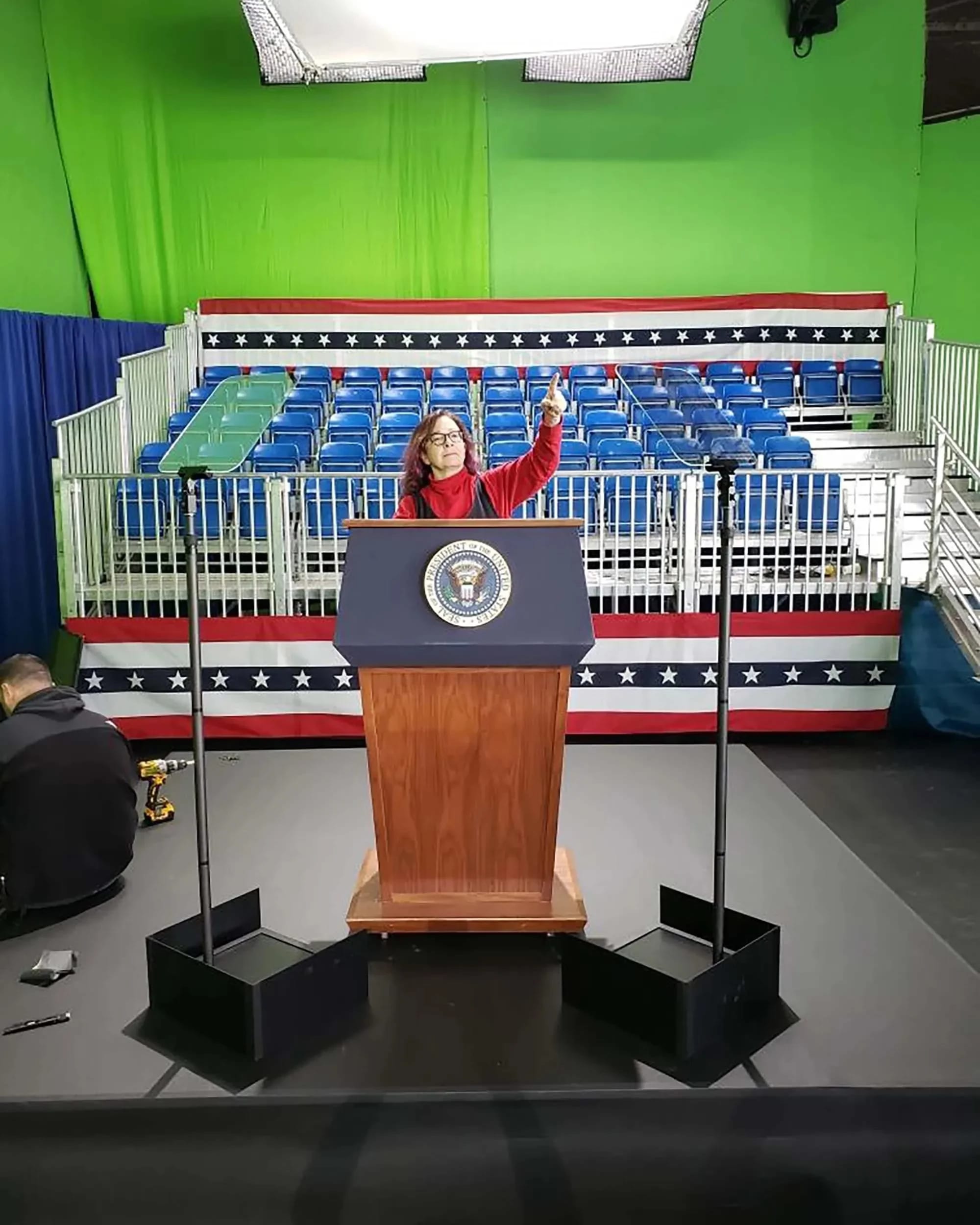 A woman in a red jacket stands at a podium with the presidential seal, facing an empty audience area with blue folding chairs arranged in rows behind her. The backdrop features a red, white, and blue design. Lighting is set up above the podium.