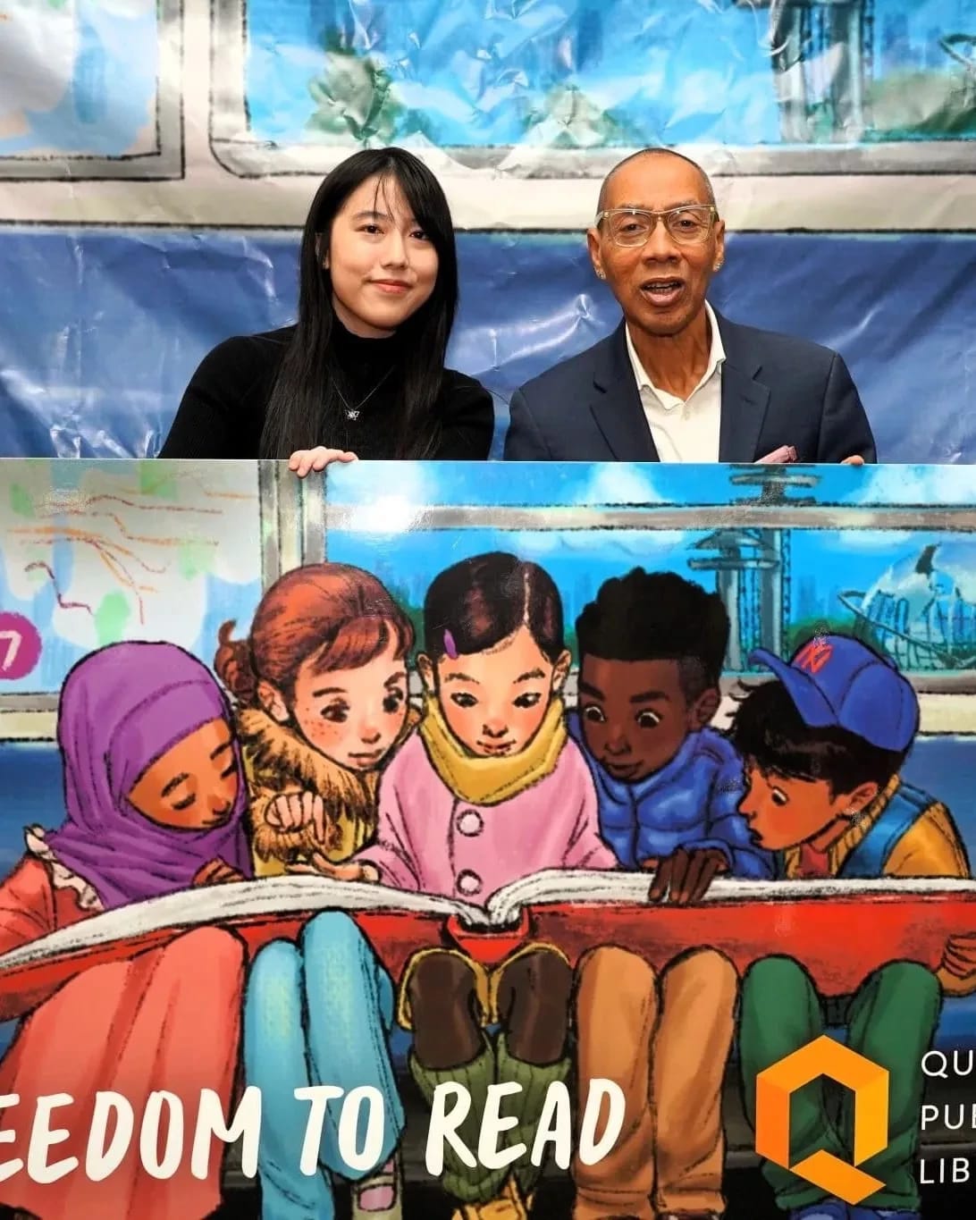 Sammi Wu sits besides a man in a suit holding a large version of a library card with an illustration of kids looking at a large red book.