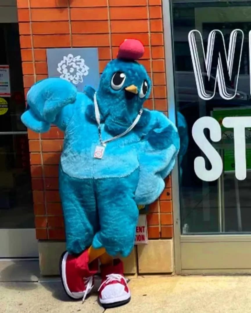 A bright blue pigeon mascot leans against a brick wall next to a window with "Welcome Students" on it in big letters.